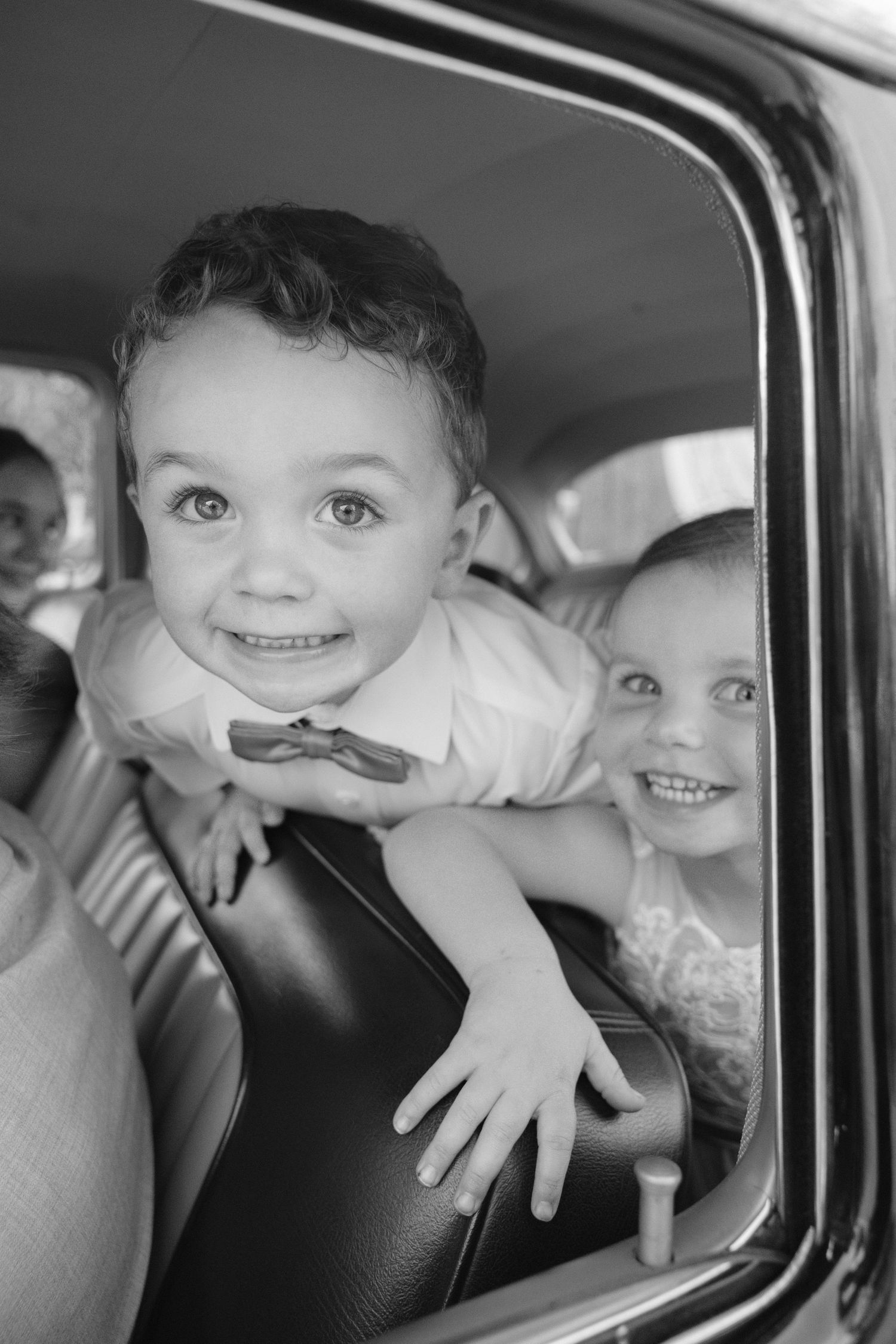 Two adorable smiling children leaning out of a vintage car window at a wedding celebration.