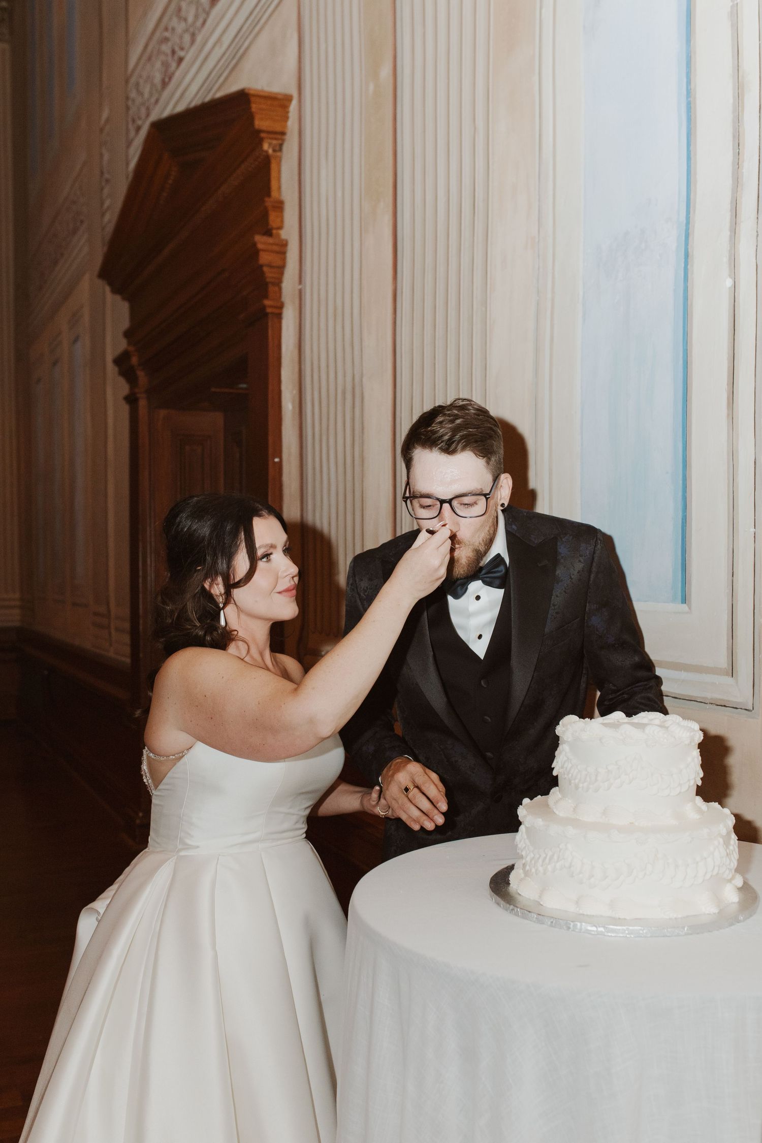 Bride feeding wedding cake to groom wearing glasses beside a two-tier white cake at elegant indoor reception.