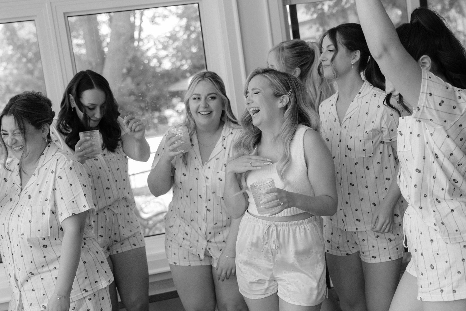 Bridesmaids in floral pajamas dancing and celebrating together by bright windows before the wedding.