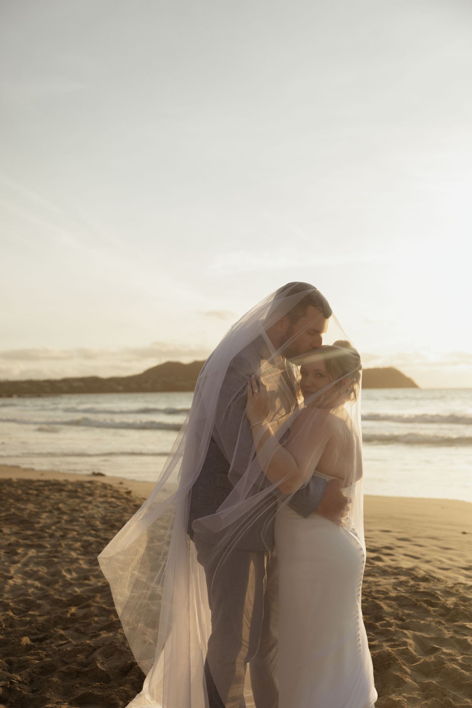 Bride and groom sharing a romantic kiss under a veil on a sandy beach at golden sunset.