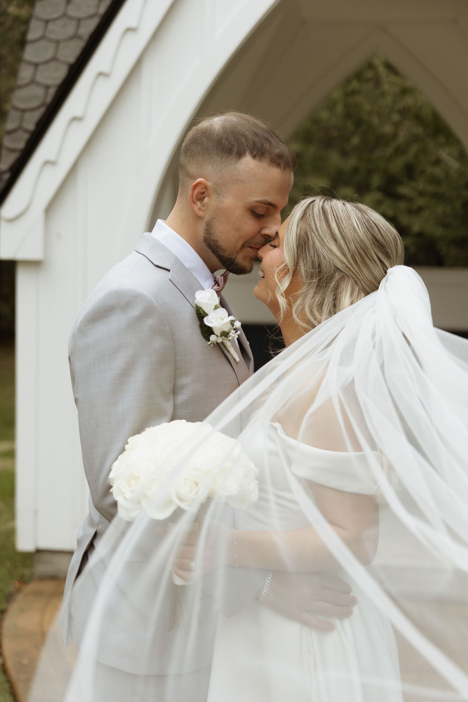 Bride and groom share a romantic moment, her veil flowing, in front of a white chapel.