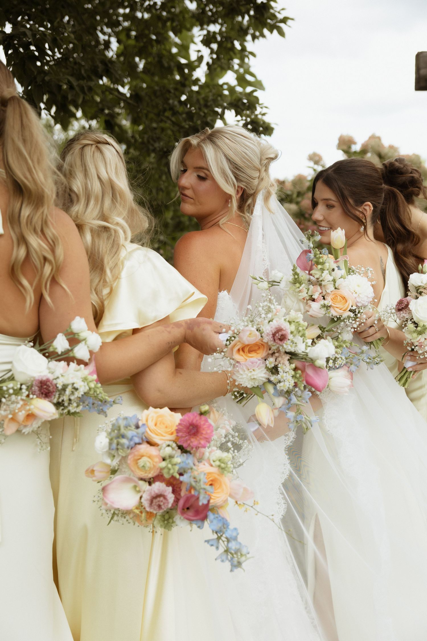 Bride in white gown and veil with bridesmaids in cream dresses holding colorful spring floral bouquets outdoors.