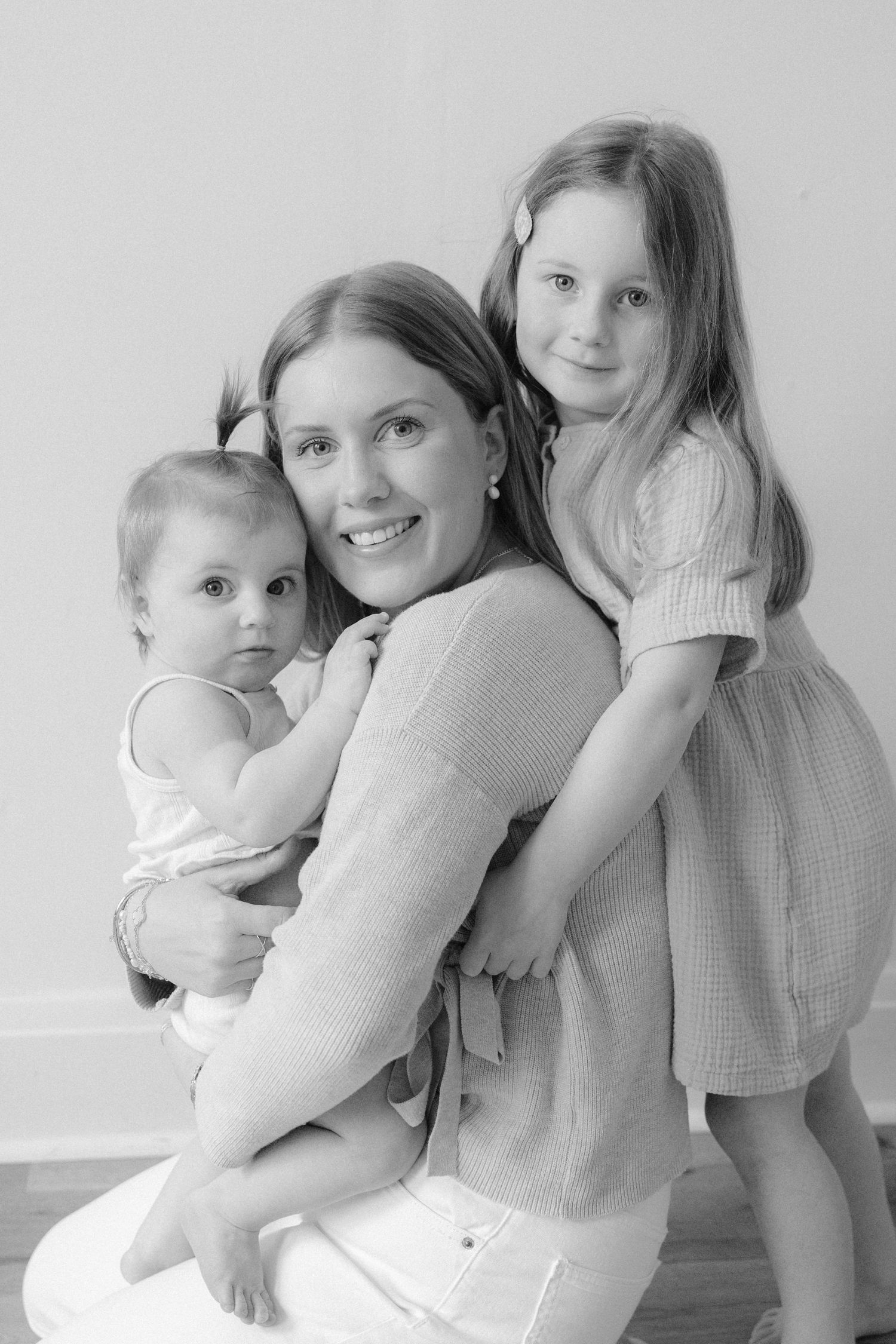 Black and white portrait of a smiling mother holding her infant and young daughter in a warm embrace.