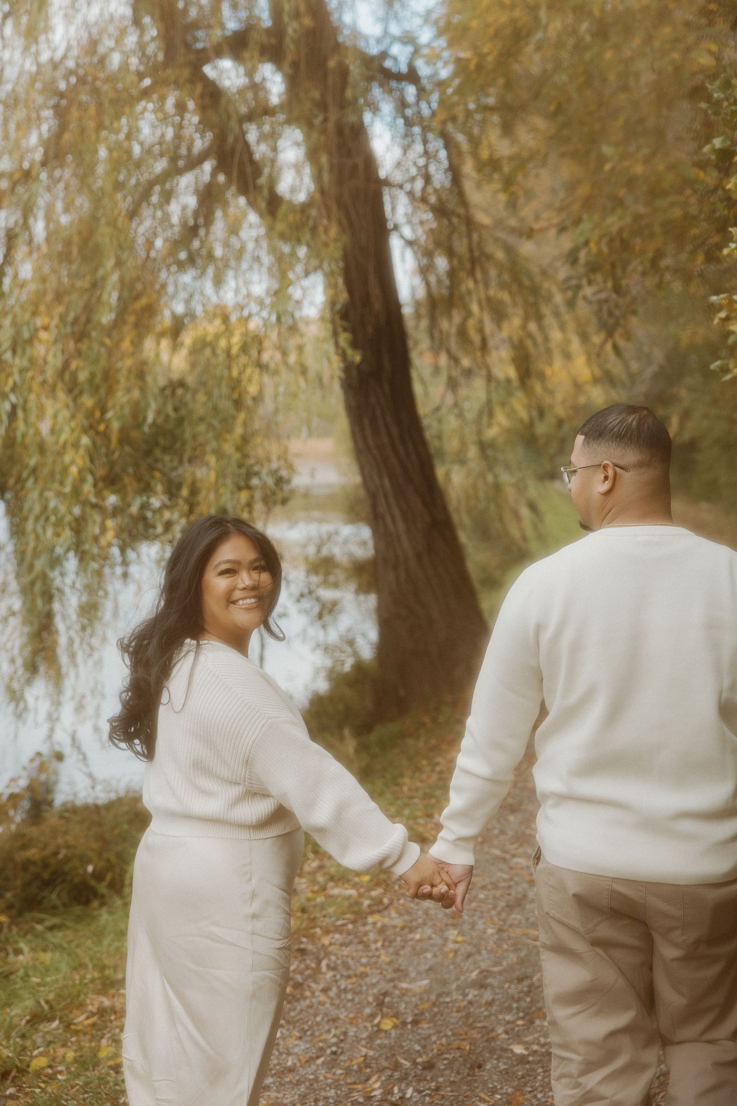 Smiling woman looks back while holding hands with partner on an autumn lakeside path.