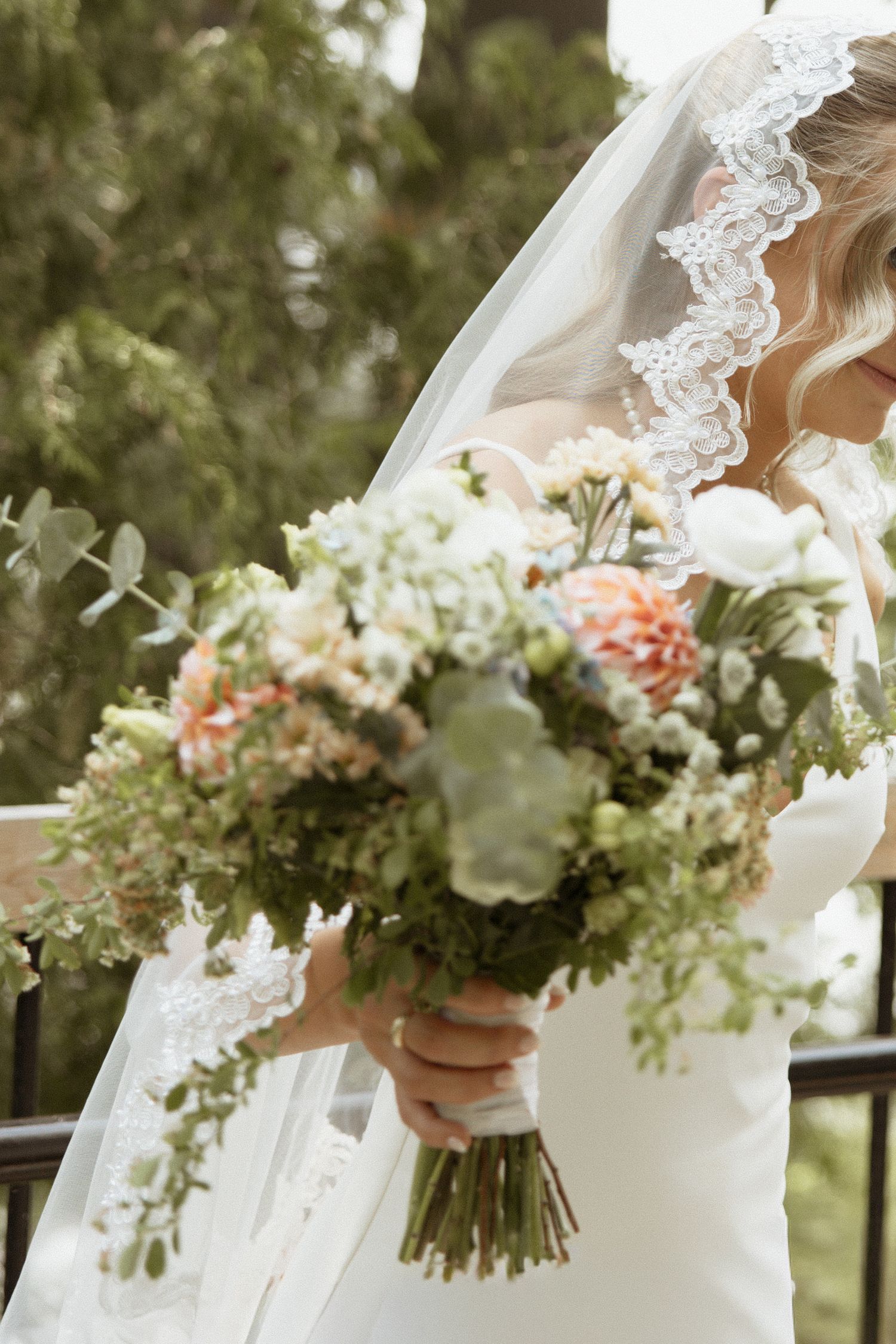 Bride in lace-trimmed veil holds lush bouquet of white and coral flowers with greenery.