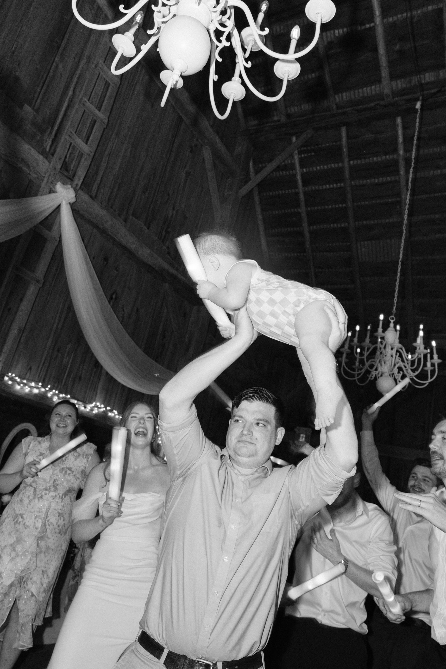 Wedding guest lifting baby overhead on crowded dance floor during festive barn reception celebration.