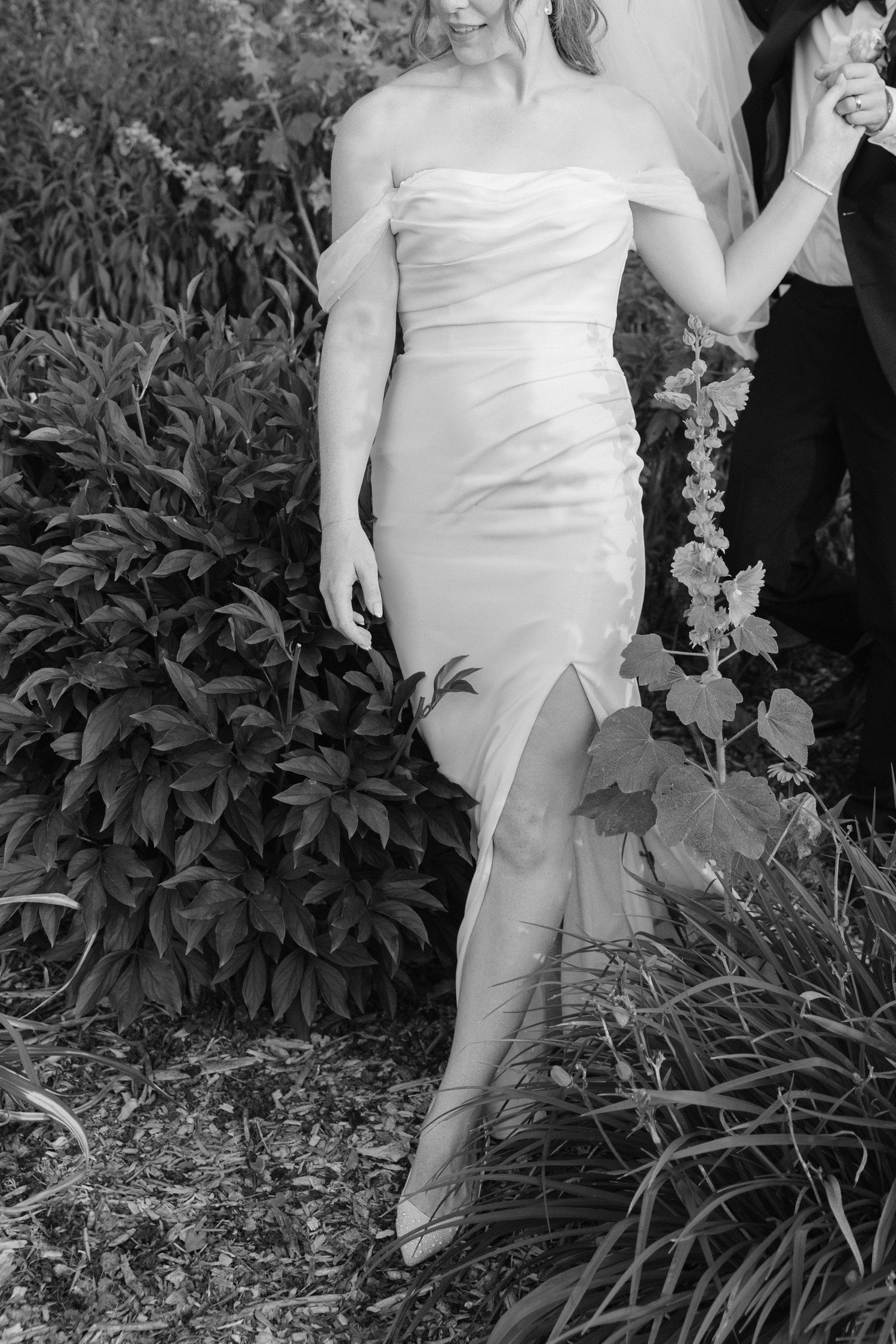 Black and white photo of bride in off-shoulder gown walking through lush garden with groom.