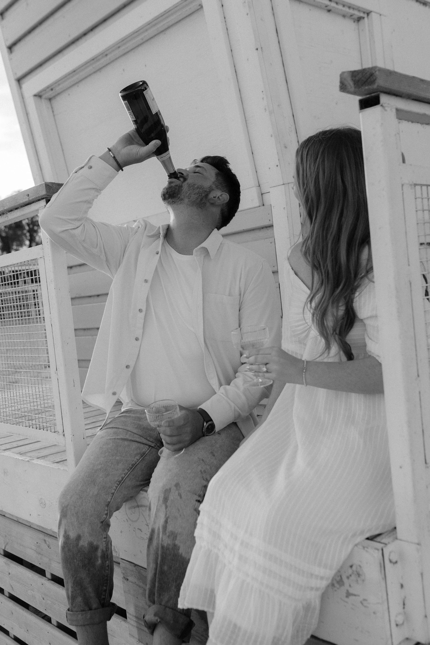 Black and white photo of man drinking from bottle beside woman on beach lifeguard stand.