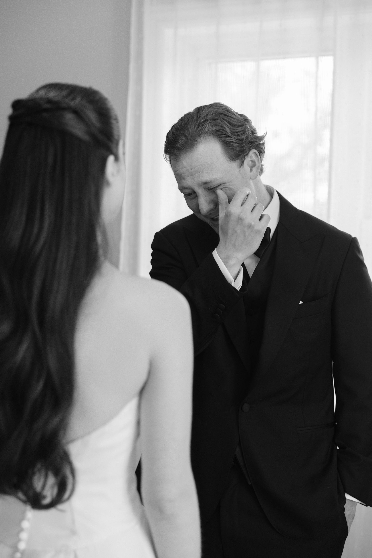 Black and white photo of emotional groom wiping tears seeing bride for first time on wedding day.