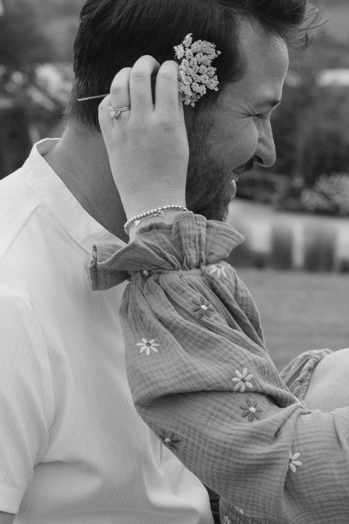 Black and white photo of a smiling bearded man holding a baby with a flower in his ear.