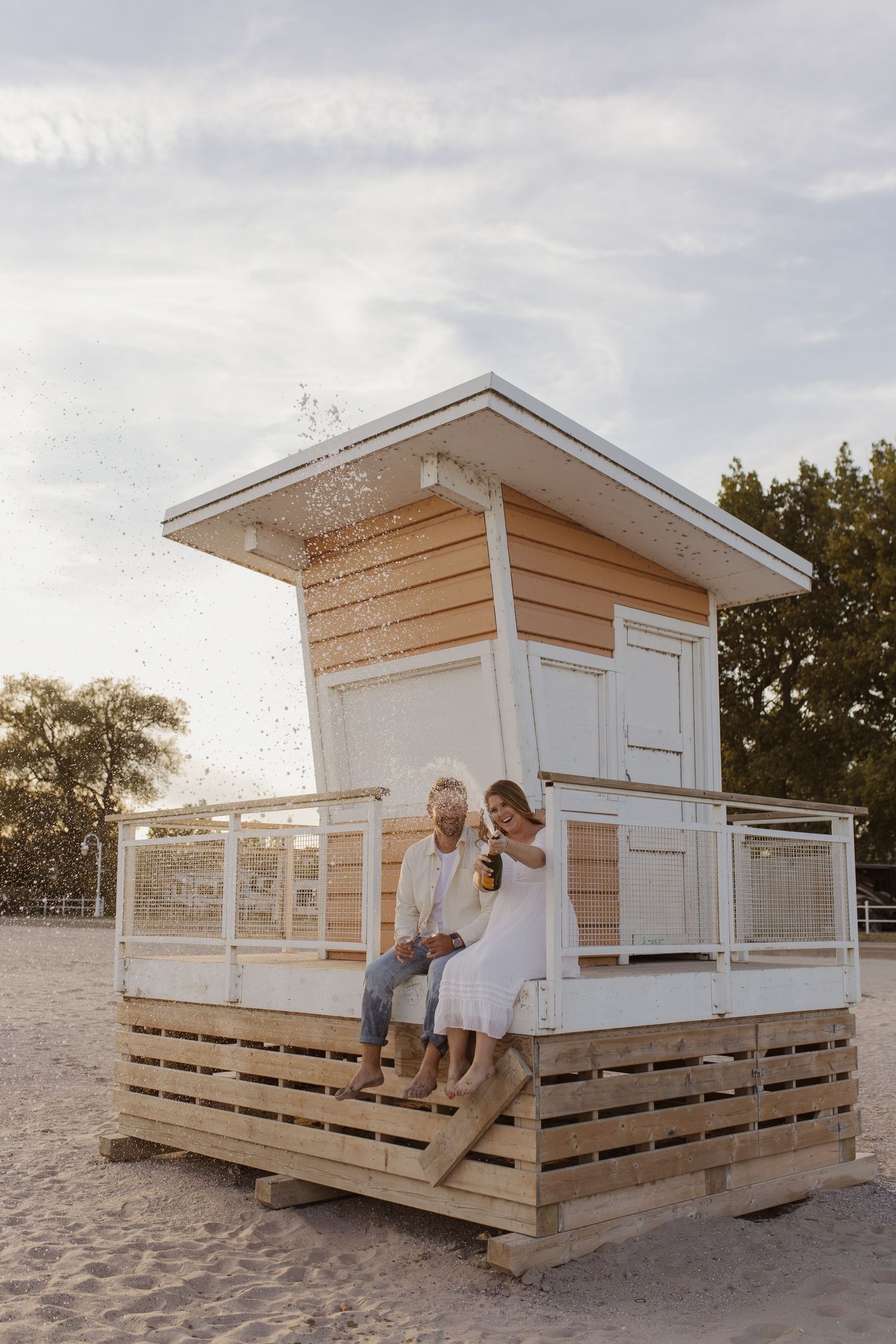 Couple popping champagne bottle on a small wooden lifeguard stand on a sandy beach.