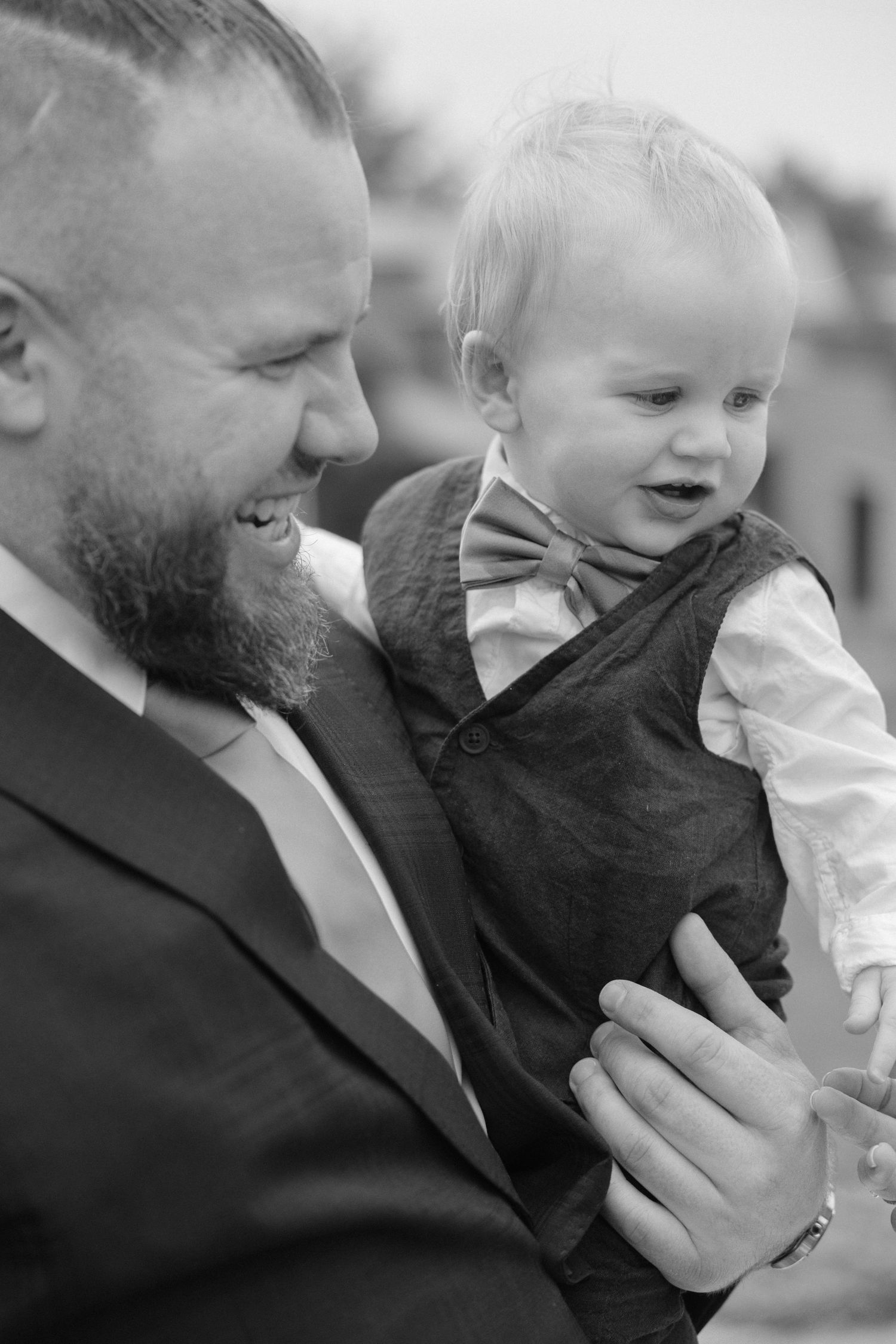 Black and white portrait of bearded groom in suit smiling while holding adorable toddler in bow tie.