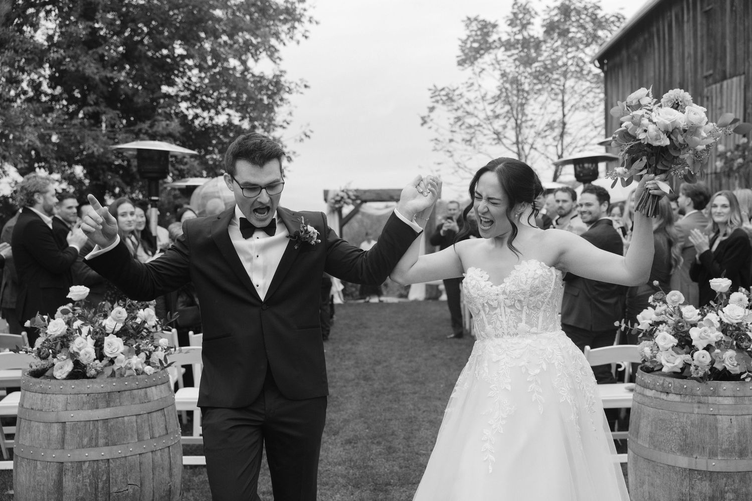 Black and white photo of joyful newlyweds raising hands walking back down aisle at rustic barn wedding.