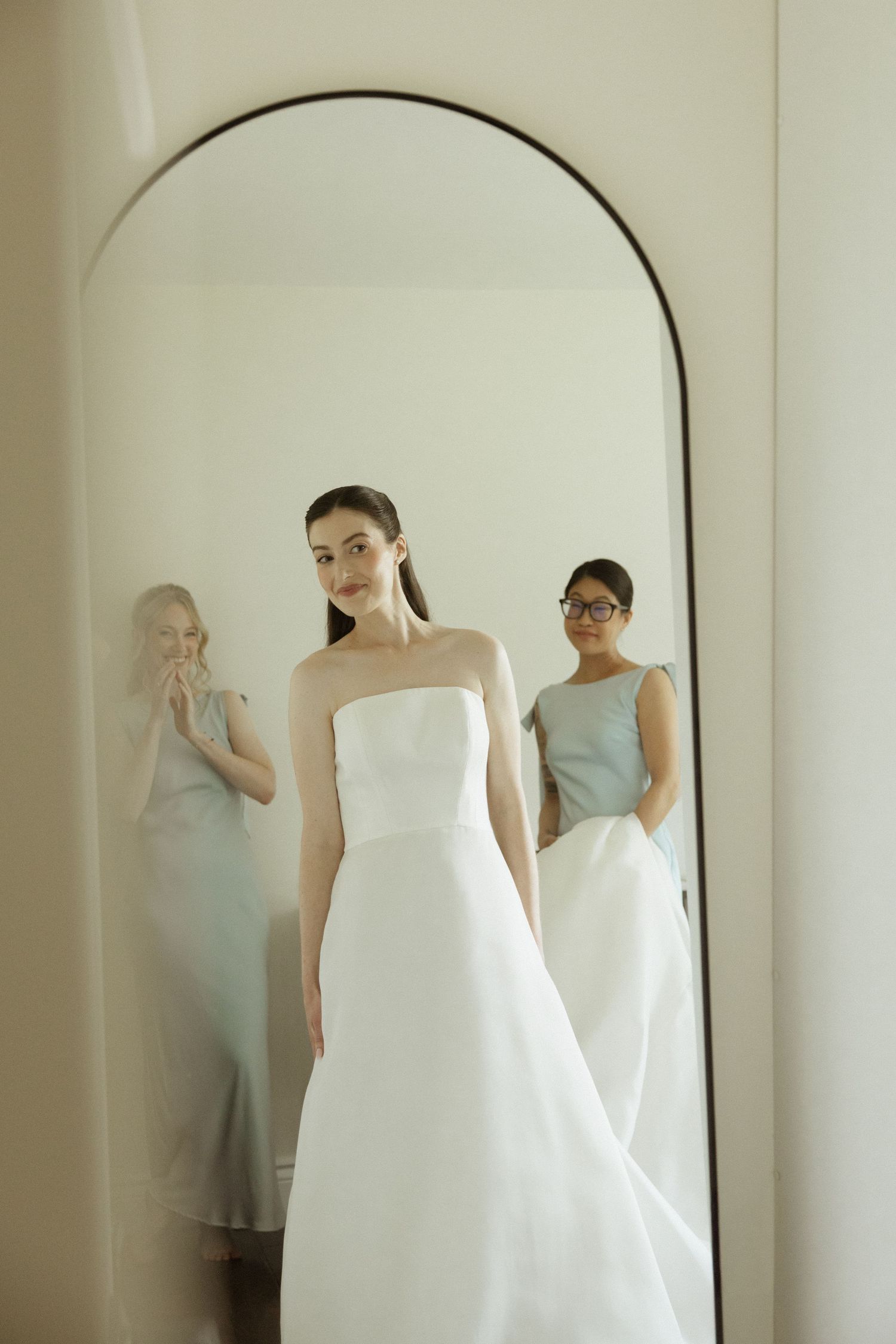 Bride in strapless white gown stands before arched mirror with bridesmaid in sage dress during wedding dress fitting.