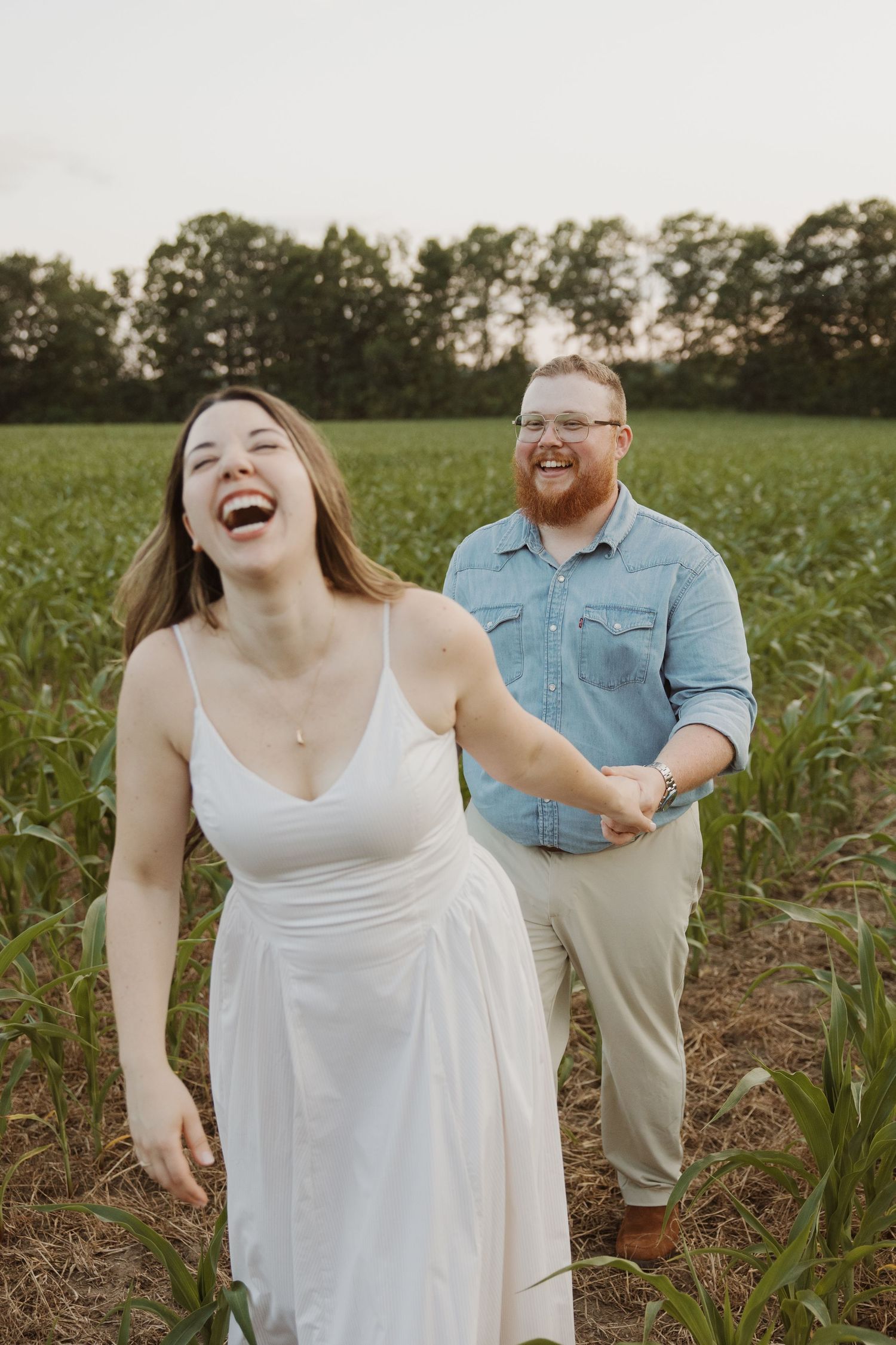 Laughing couple holding hands in a green cornfield at dusk.
