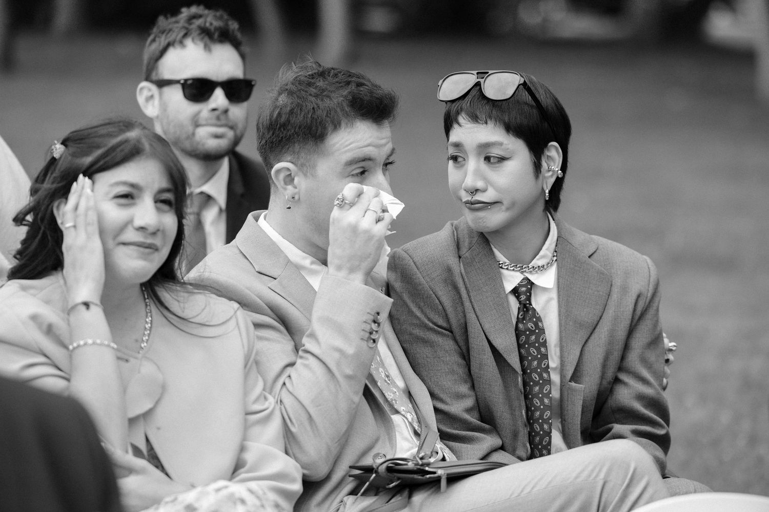 Black and white photo of emotional wedding guests, one dabbing tears with tissue during outdoor ceremony.