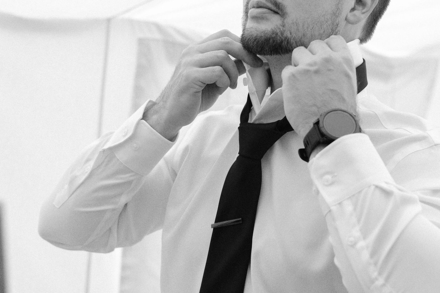 Black and white photo of a groom adjusting his tie while getting ready for his wedding.