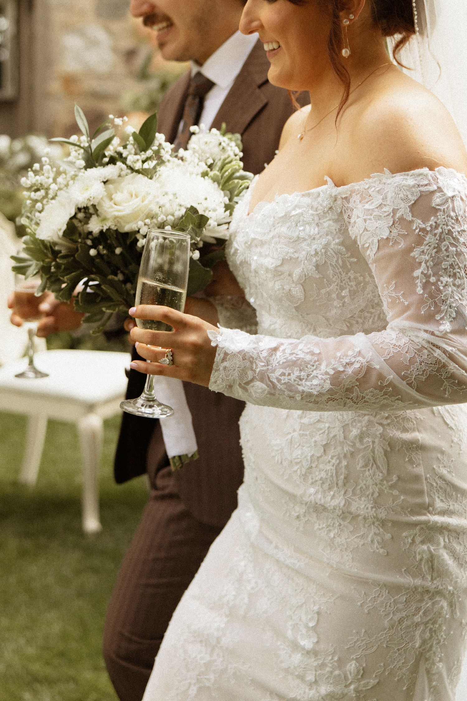 Bride in lace off-shoulder gown holds champagne flute and white floral bouquet beside groom.