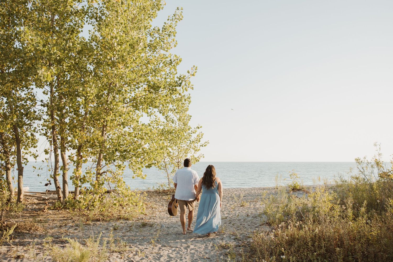 Couple holding hands walking toward beach through golden trees in warm sunlight.