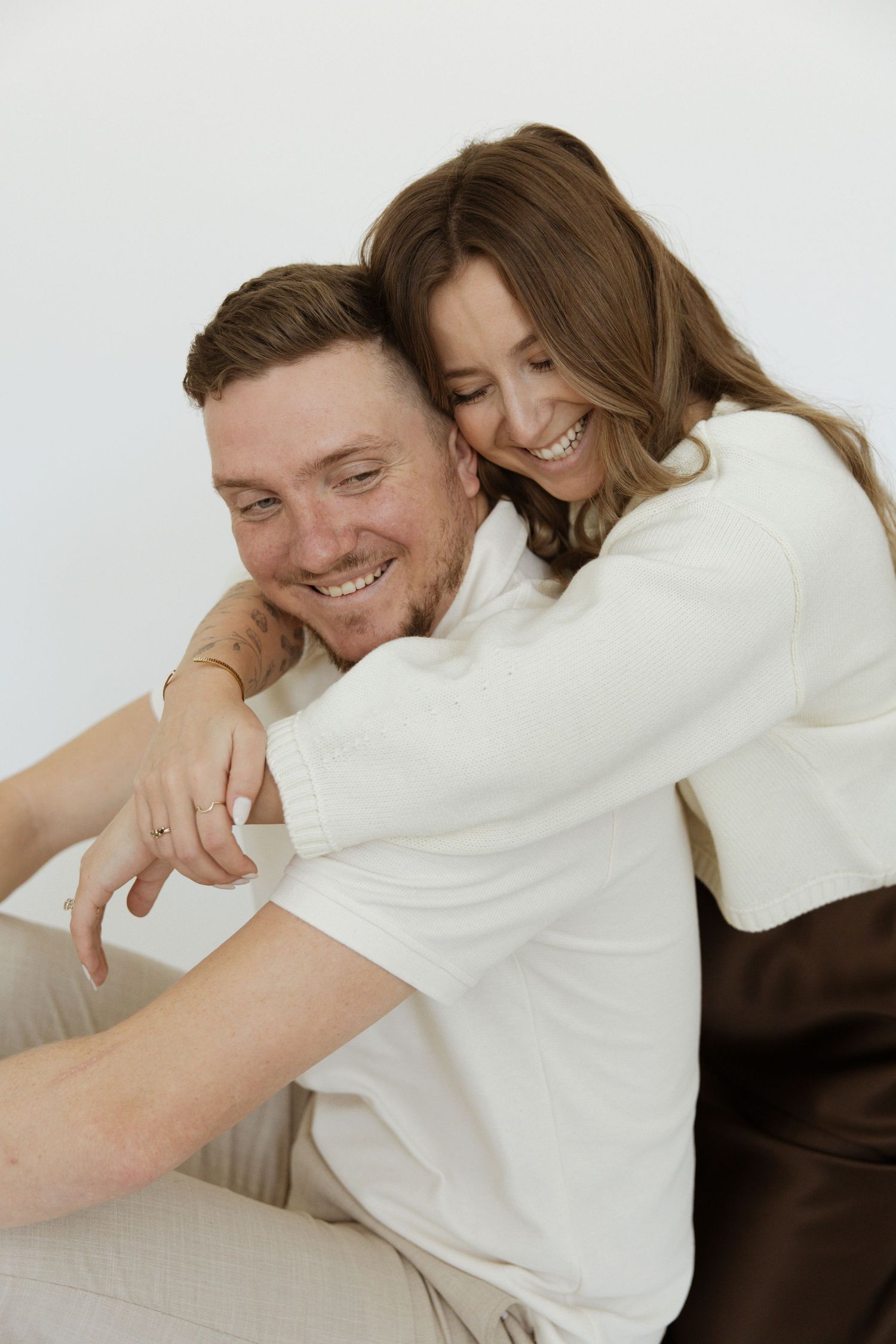 Smiling woman embracing man from behind as they sit together laughing against a white background.