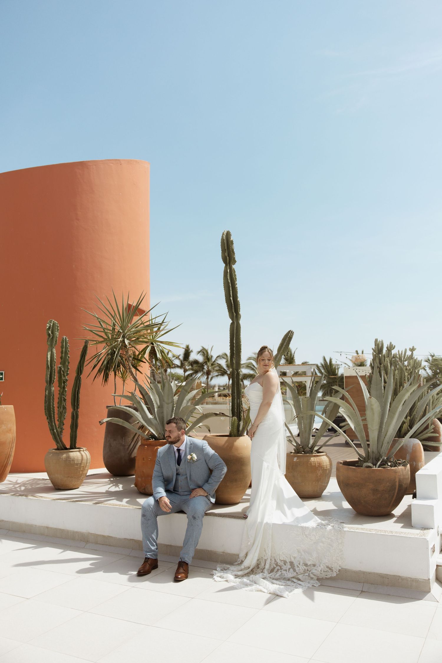 Bride and groom pose on a white terrace surrounded by cacti and agave plants with a terracotta wall in Cabo.