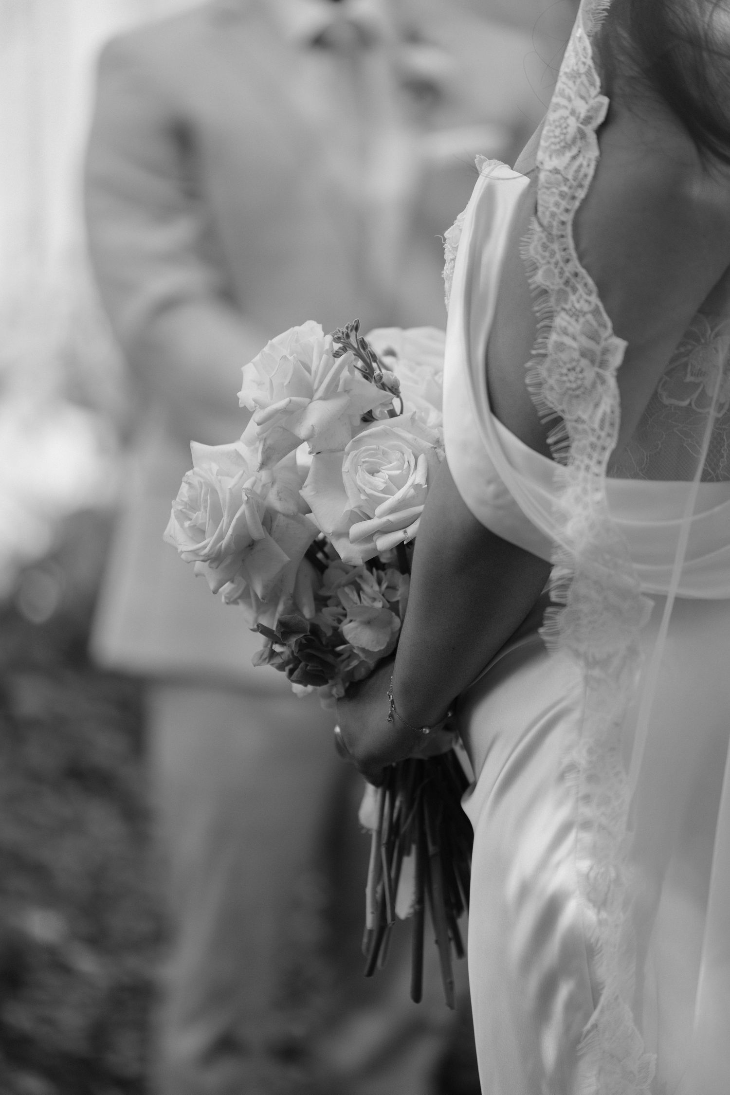 Black and white wedding photo of bride holding rose bouquet in lace gown with groom standing behind her.