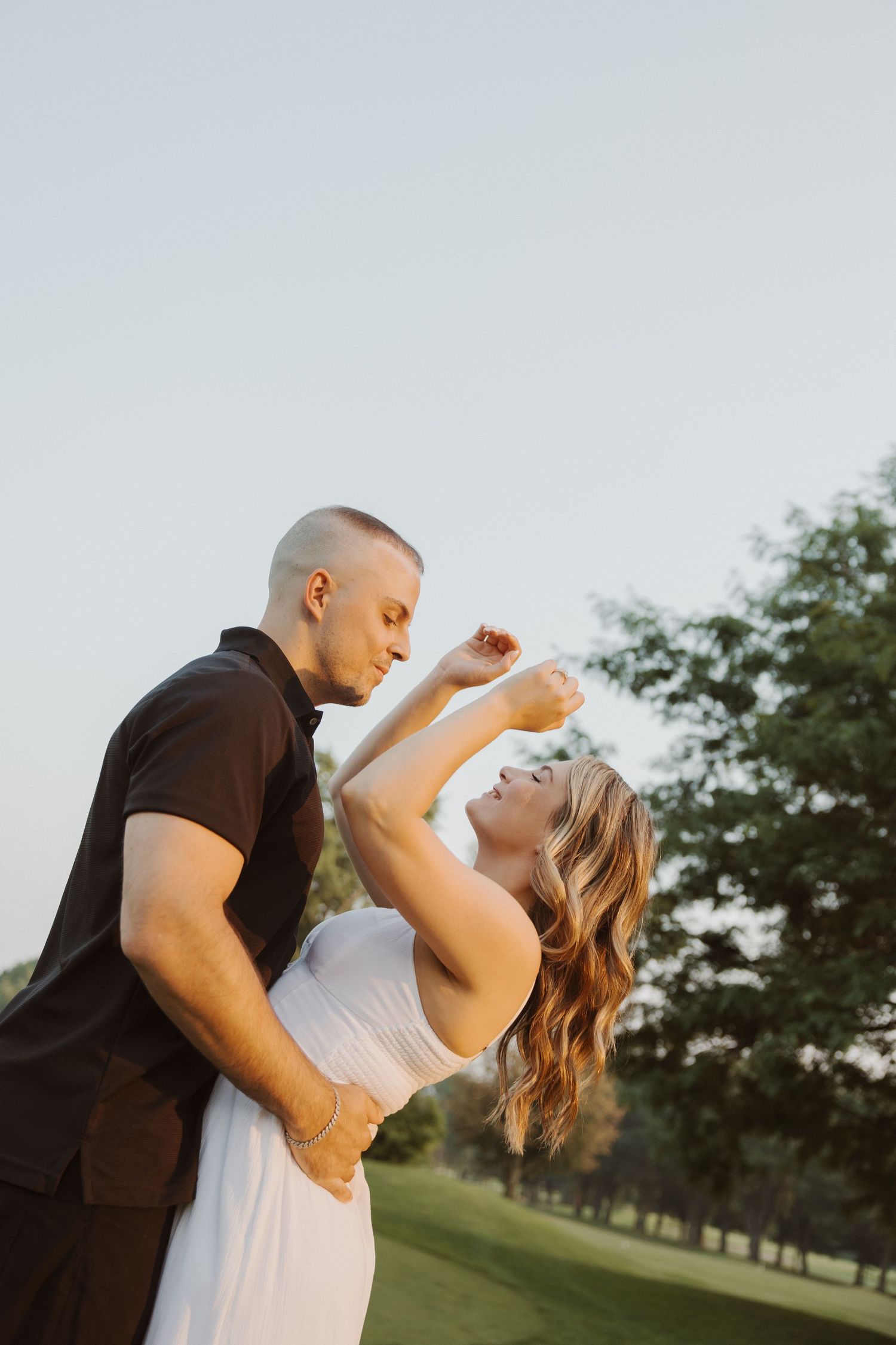 Couple dancing outdoors at sunset, woman in white dress laughing as man in black shirt holds her close.
