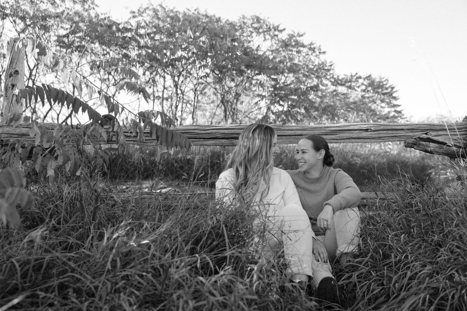Black and white photo of two women sitting in tall grass near a wooden fence surrounded by trees.