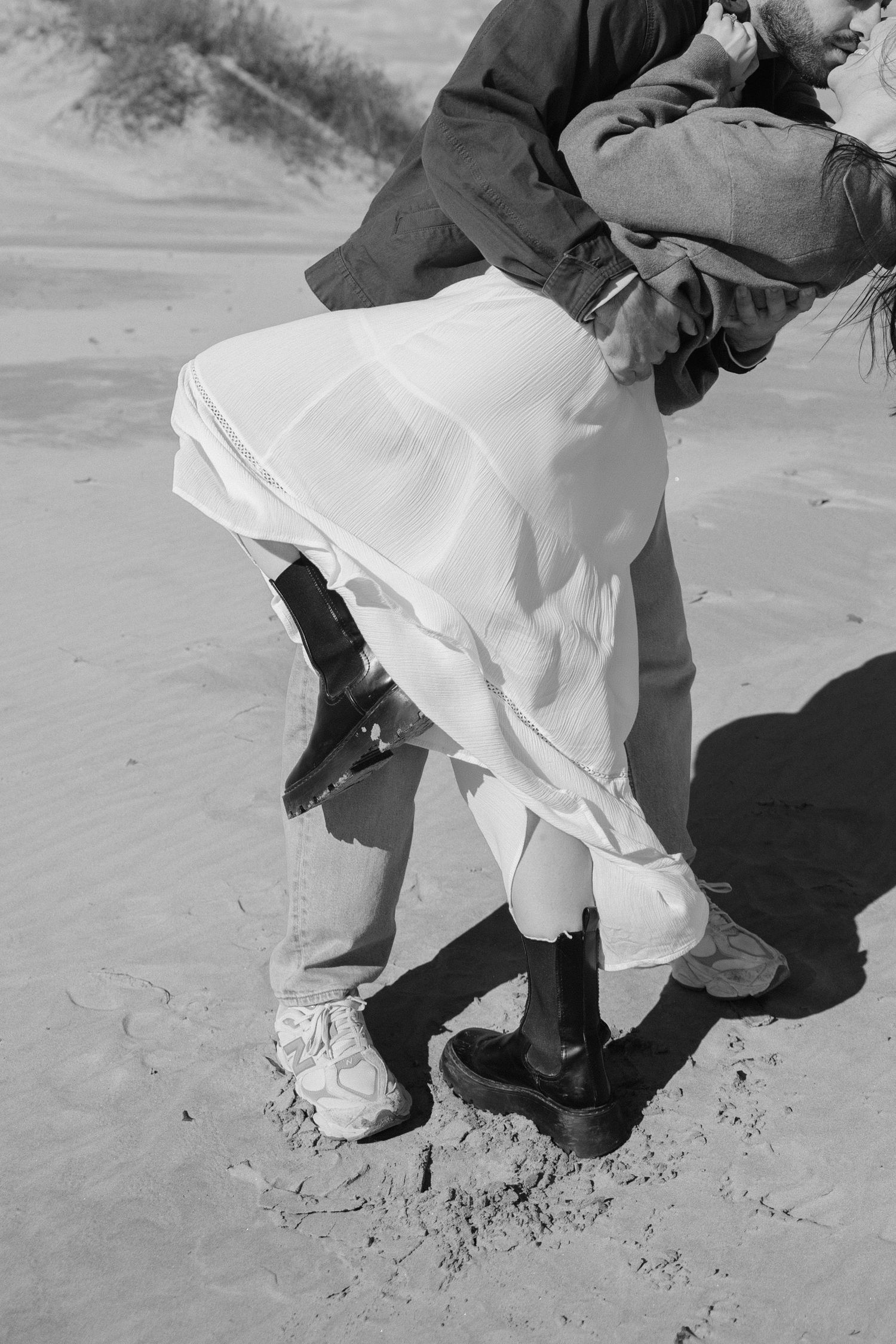 Black and white close-up of couple embracing on sandy beach, woman wearing white dress and black boots.