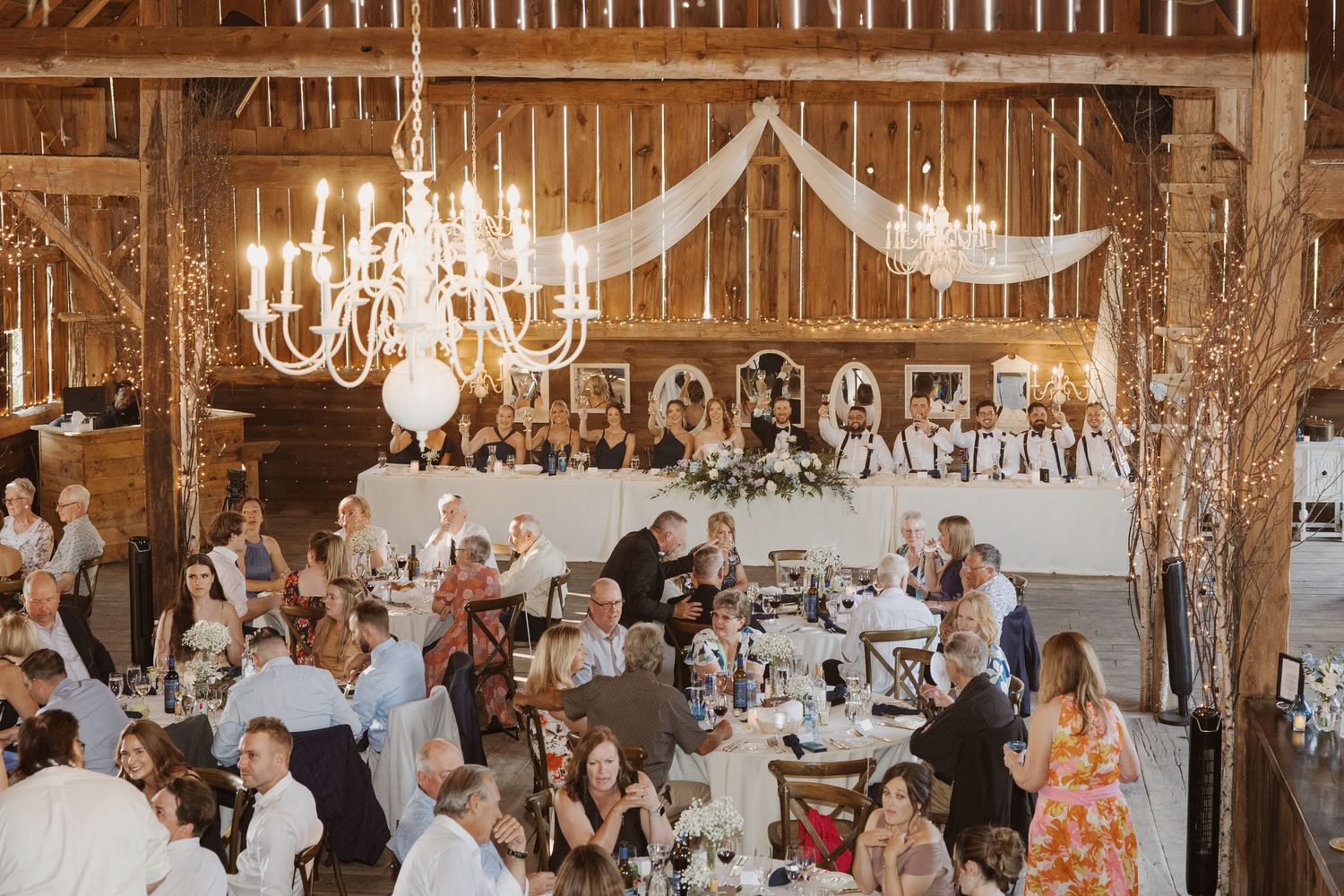 Elegant barn wedding reception with chandeliers, draped fabric, and guests dining at round tables.