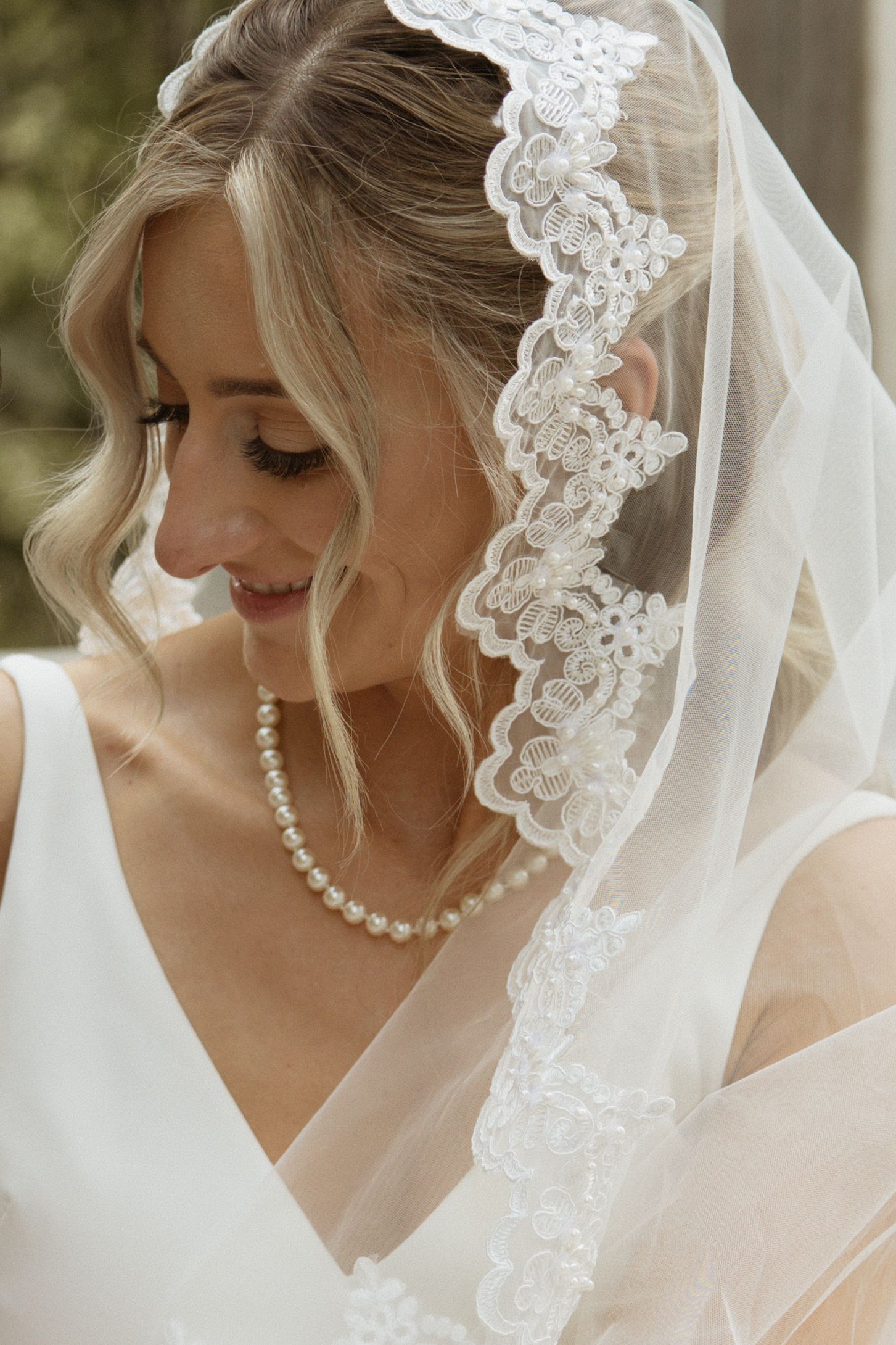 Beautiful bride wearing lace-trimmed veil and pearl necklace on wedding day.