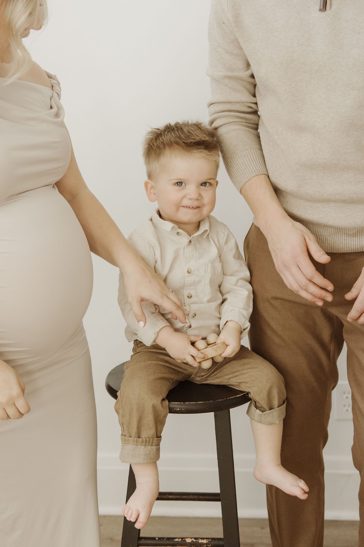 Smiling toddler boy in linen outfit sitting on a stool between his pregnant mother and father.