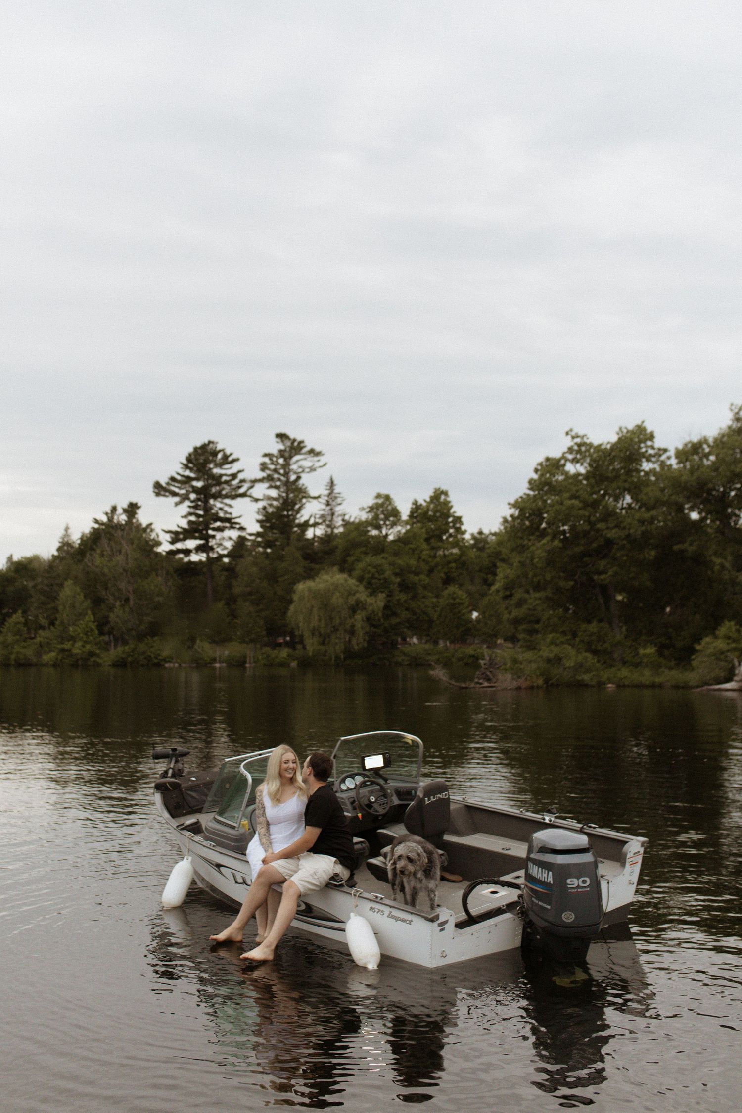 Couple relaxing on a motorboat on a calm lake surrounded by lush green trees on an overcast day.