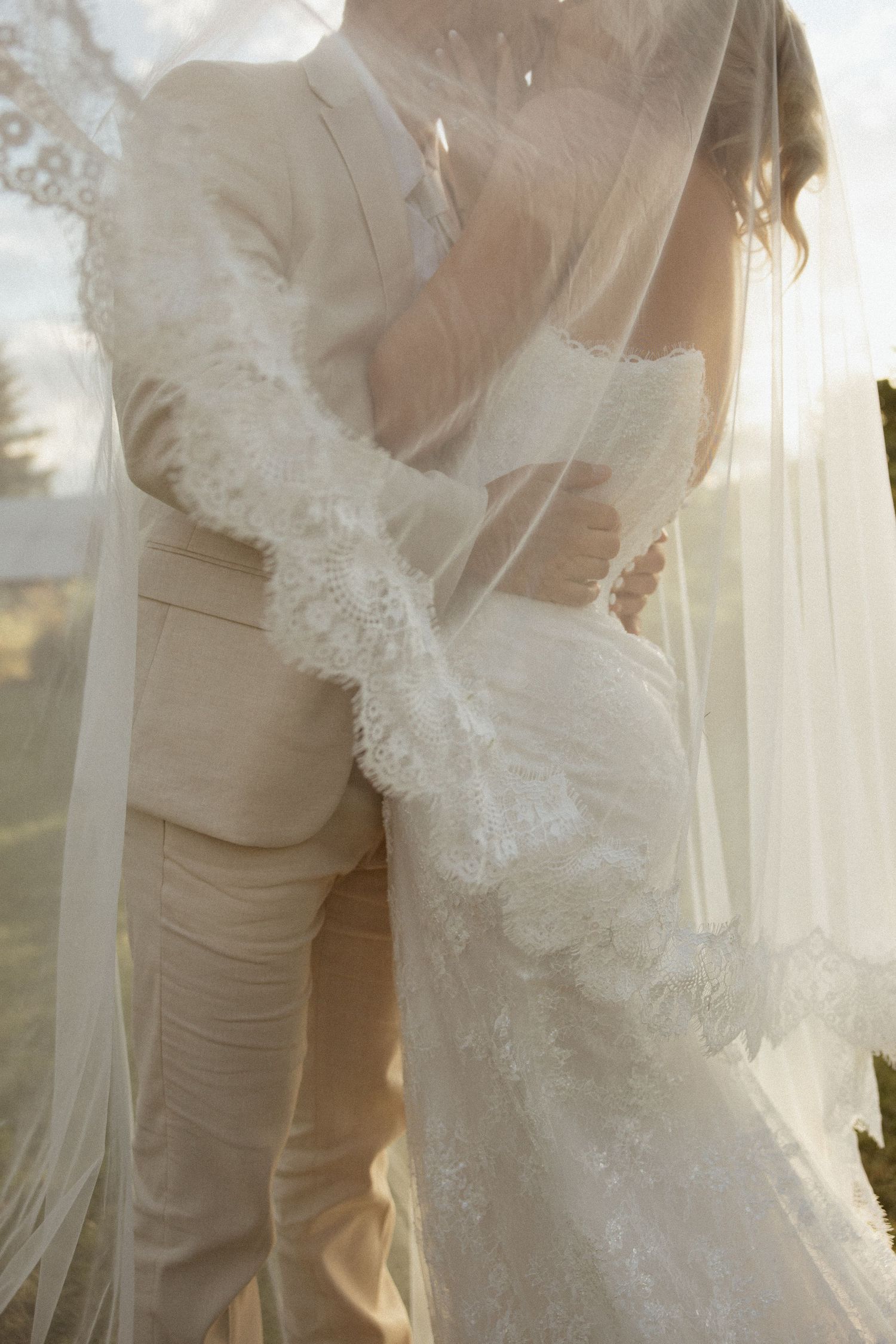 Bride and groom embracing under a flowing lace wedding veil in warm golden light.