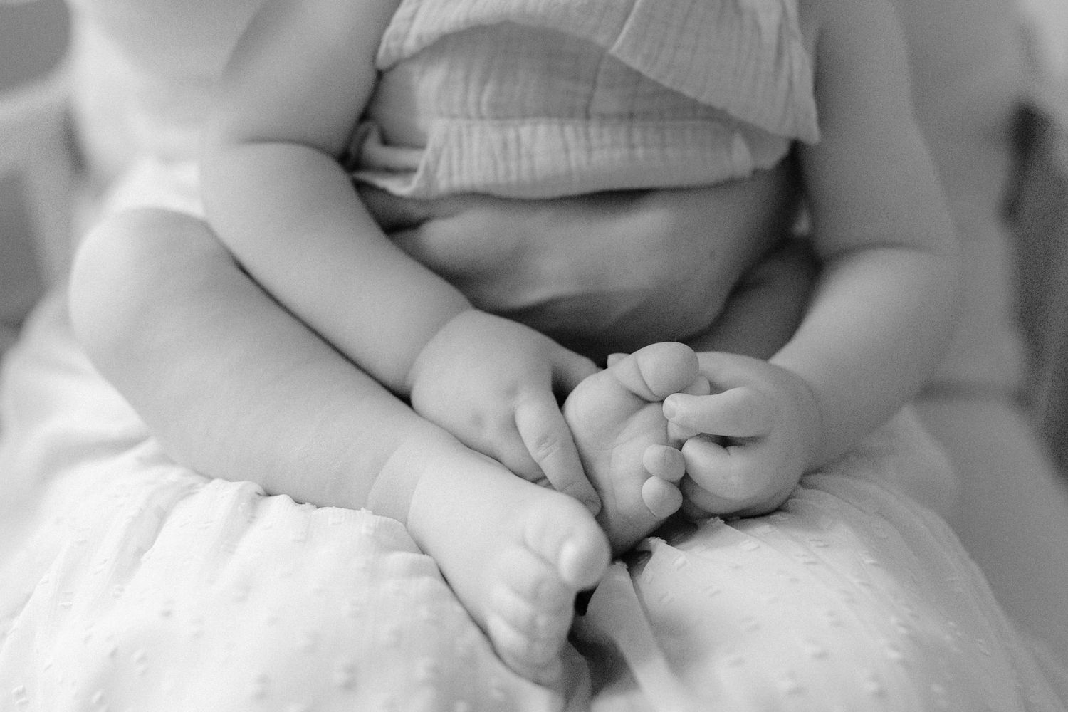 Black and white close-up of a baby holding their own tiny feet while sitting on a soft white blanket.