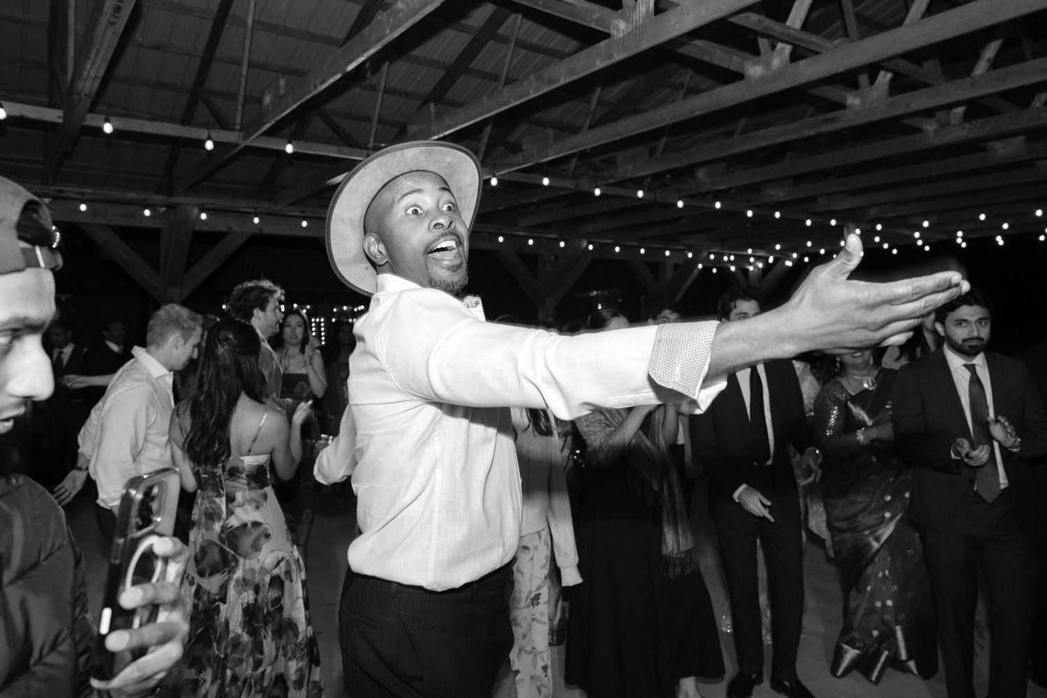 Black and white photo of a joyful guest dancing energetically at a wedding reception under string lights.