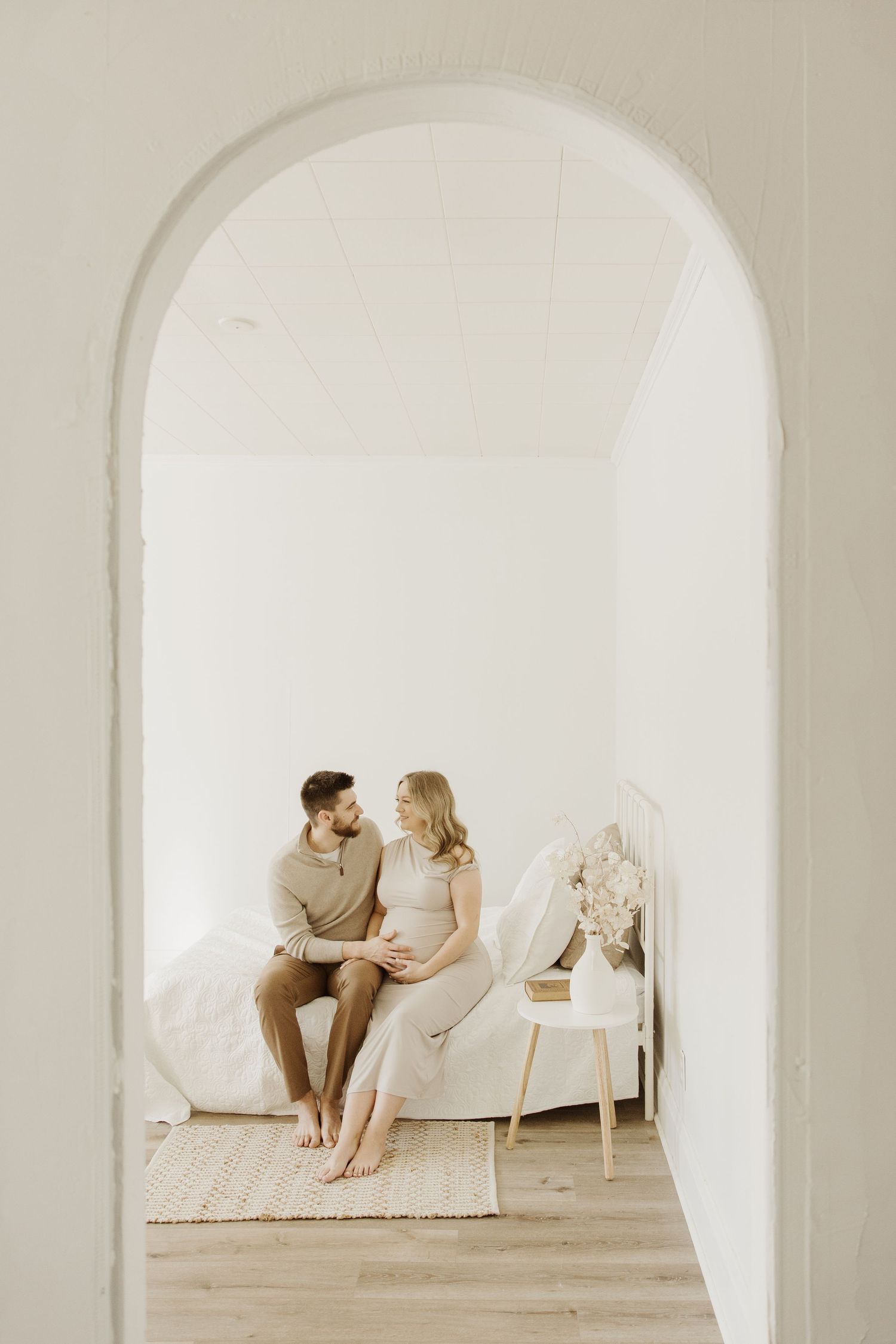 Expectant couple sitting on a bed together viewed through a white archway in a bright minimal room.