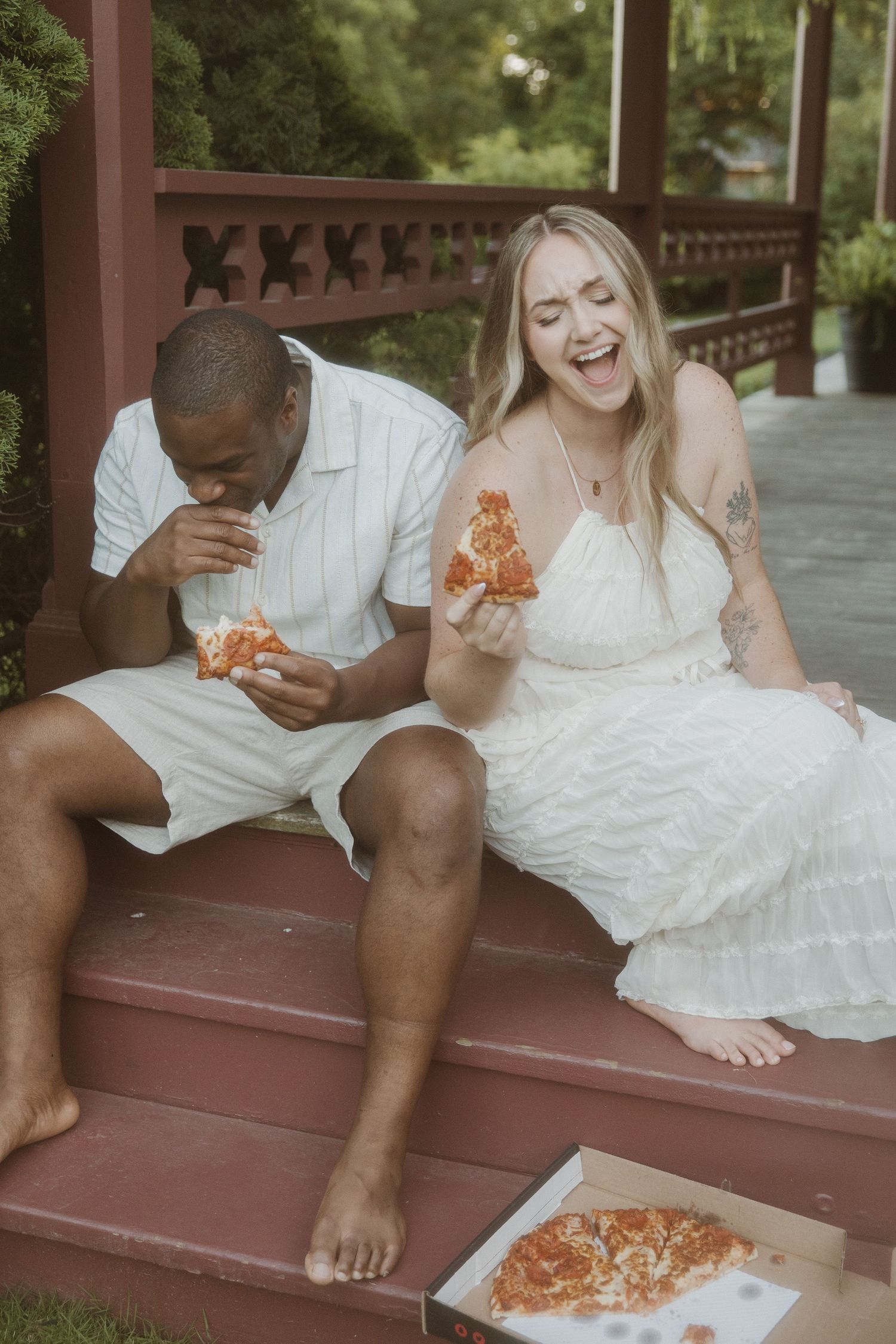 Couple in white outfits laughing and eating pizza on porch steps outdoors.