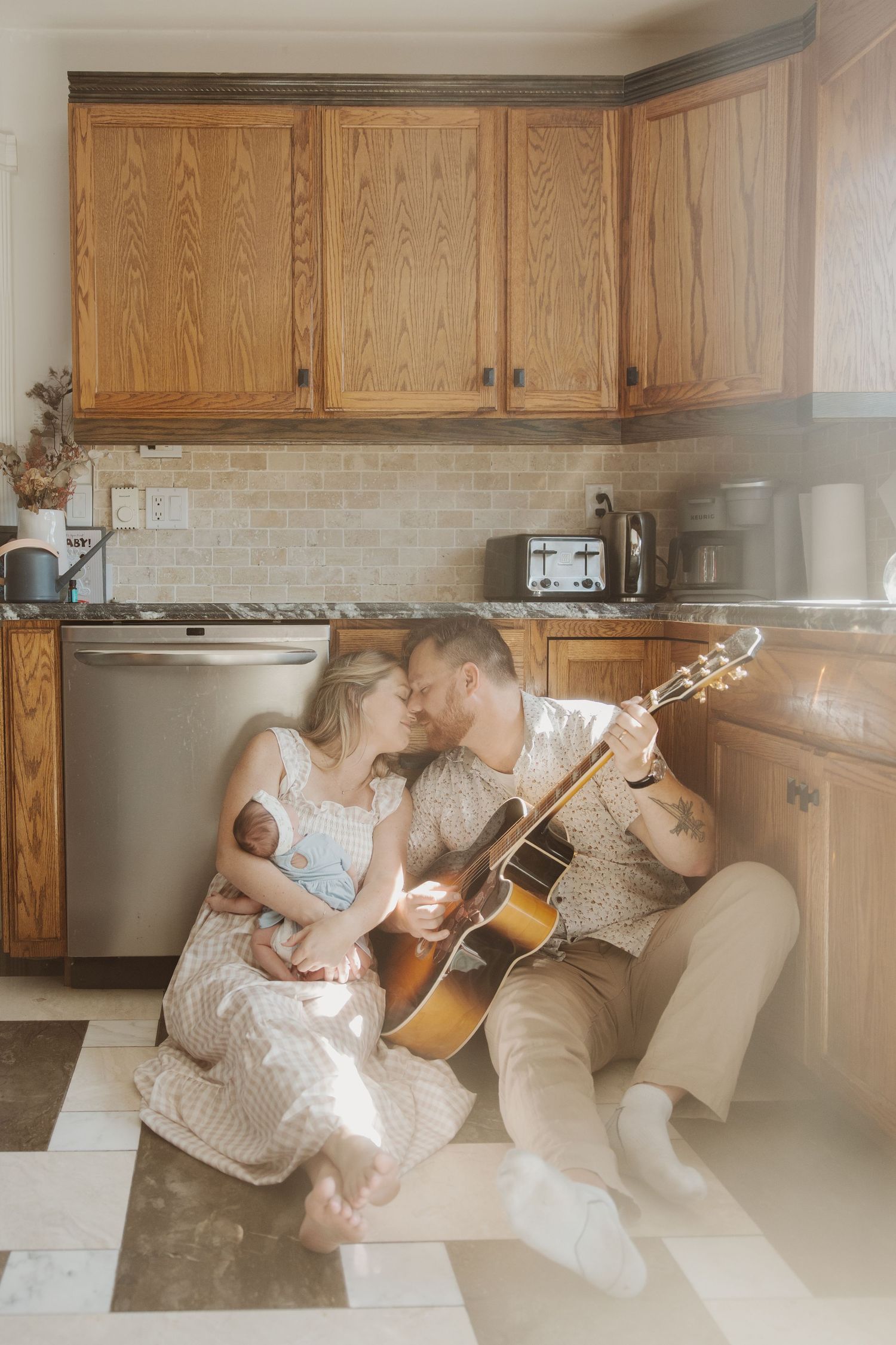 Couple sharing a kiss on kitchen floor, man playing guitar while woman holds newborn baby.