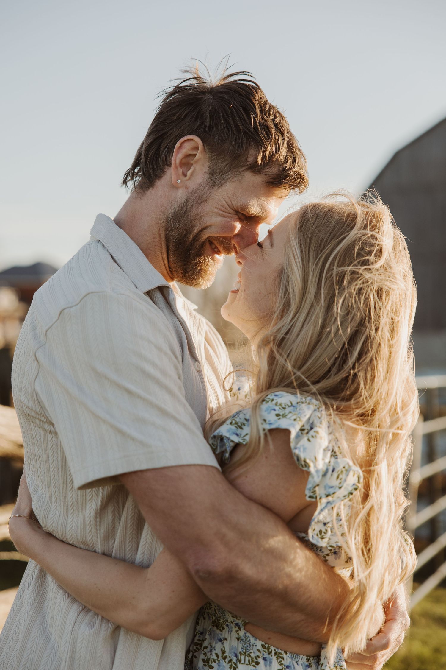 Romantic couple embracing and almost kissing outdoors at golden hour near a white fence.