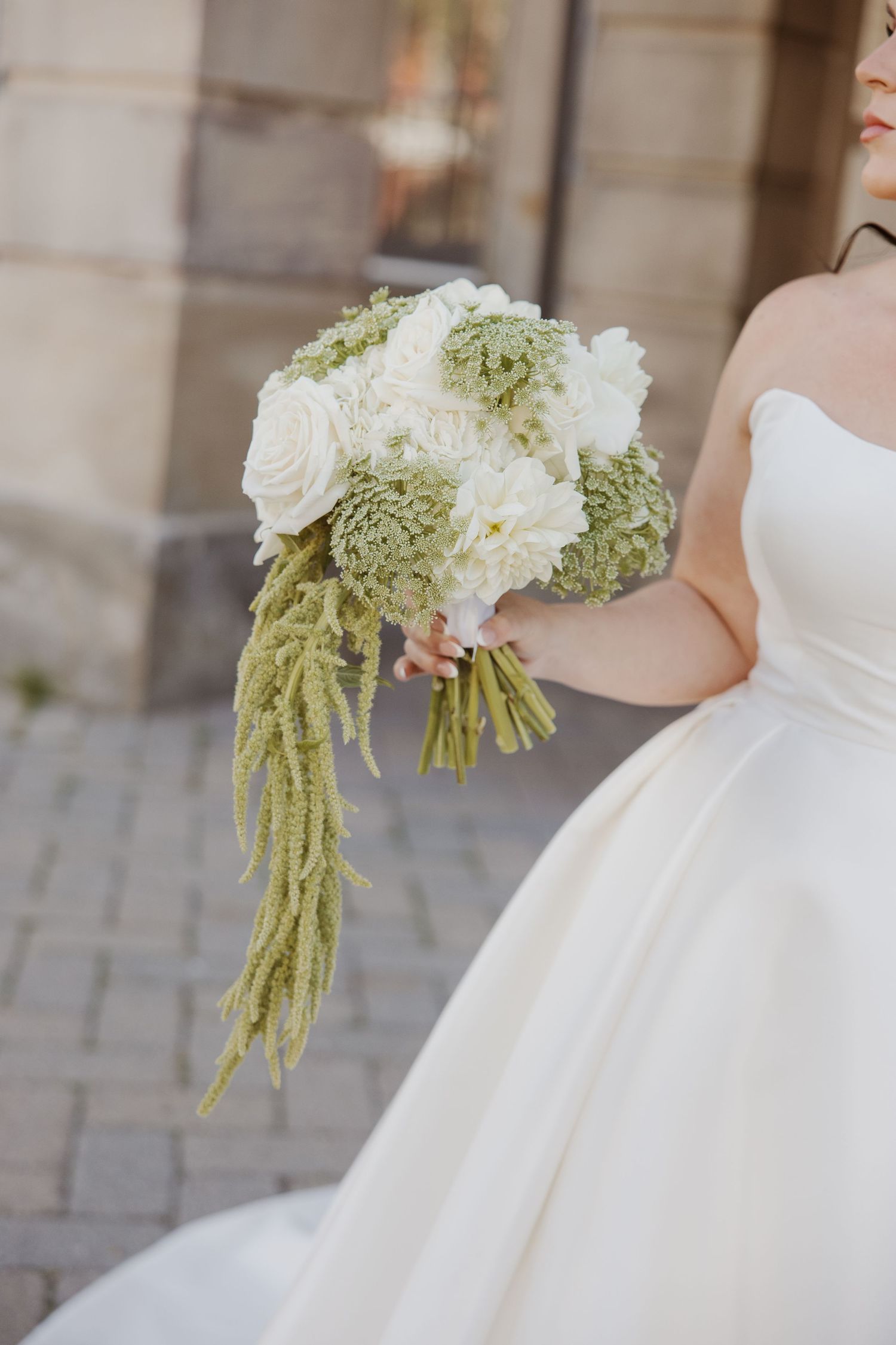 Bride in strapless white gown holds a bouquet of white roses and green succulents with trailing amaranthus.