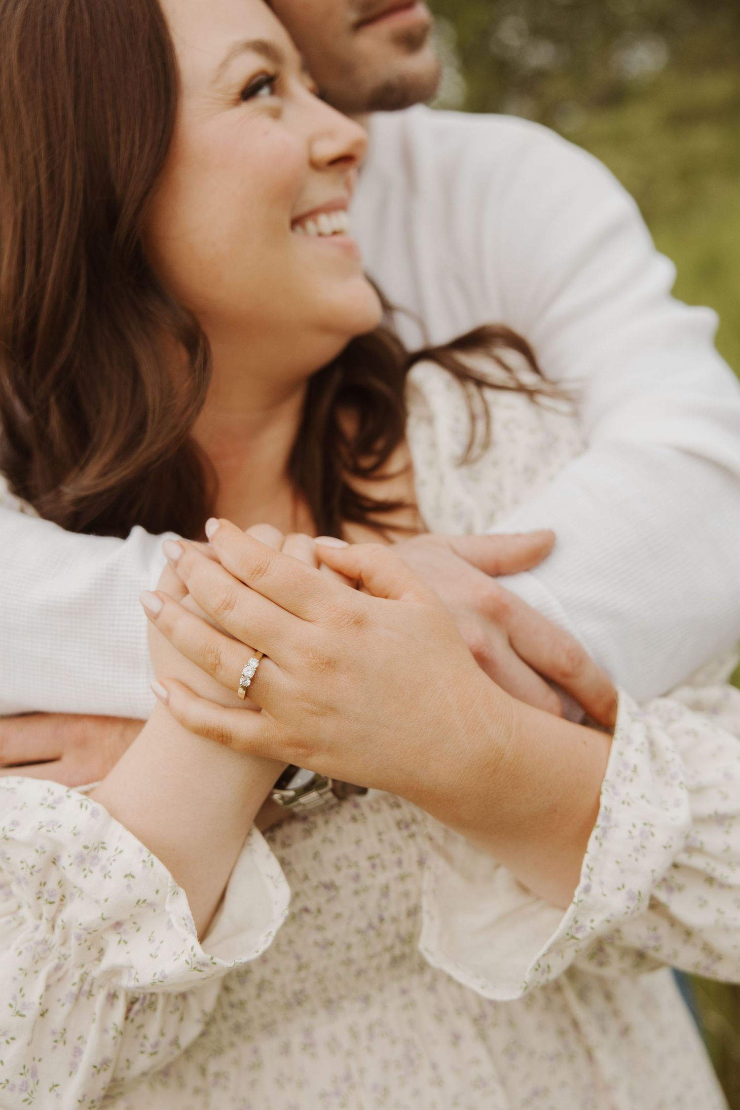Close-up of woman smiling with engagement ring visible as partner wraps arms around her from behind.