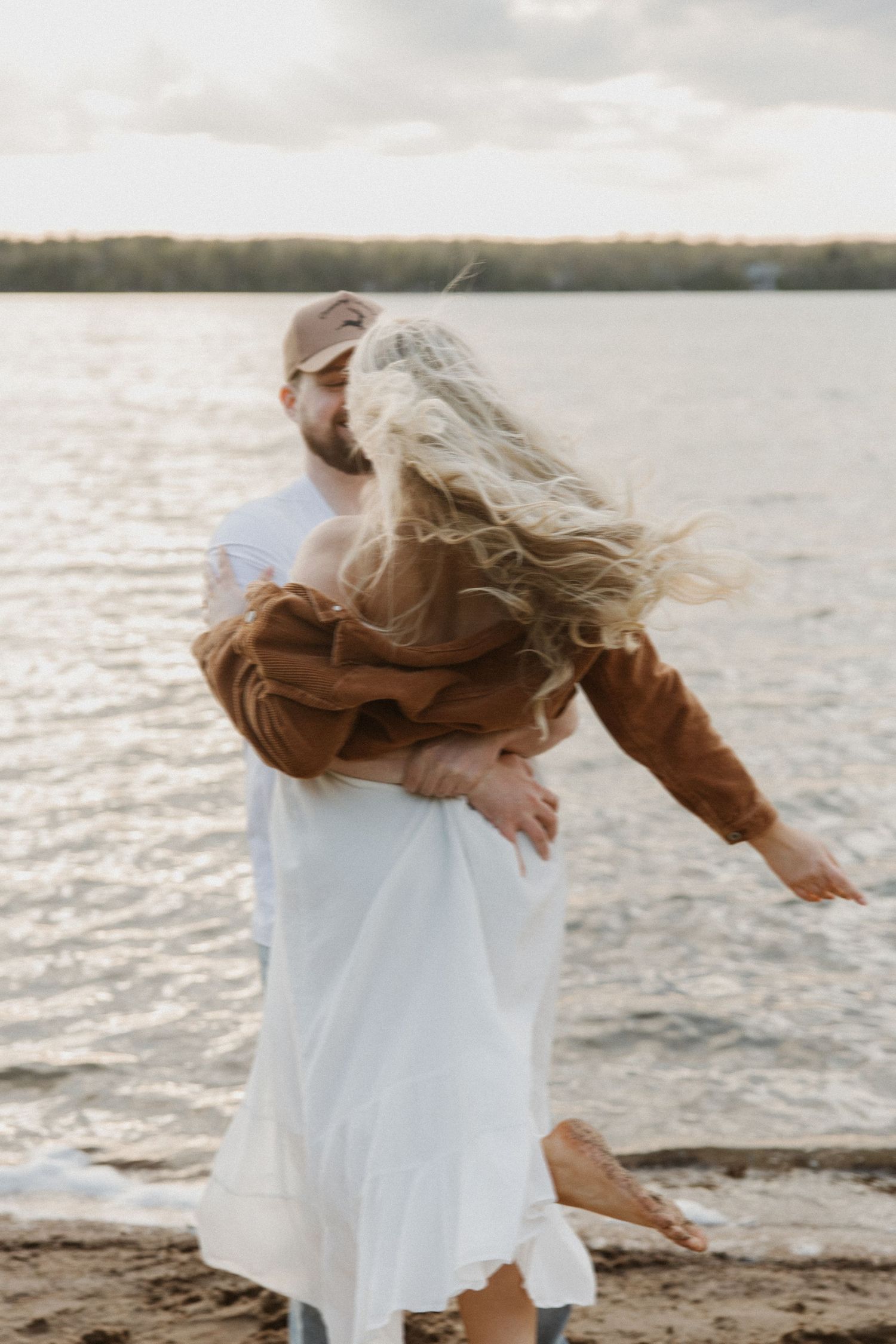 Man lifting woman in brown jacket on sandy beach at sunset.