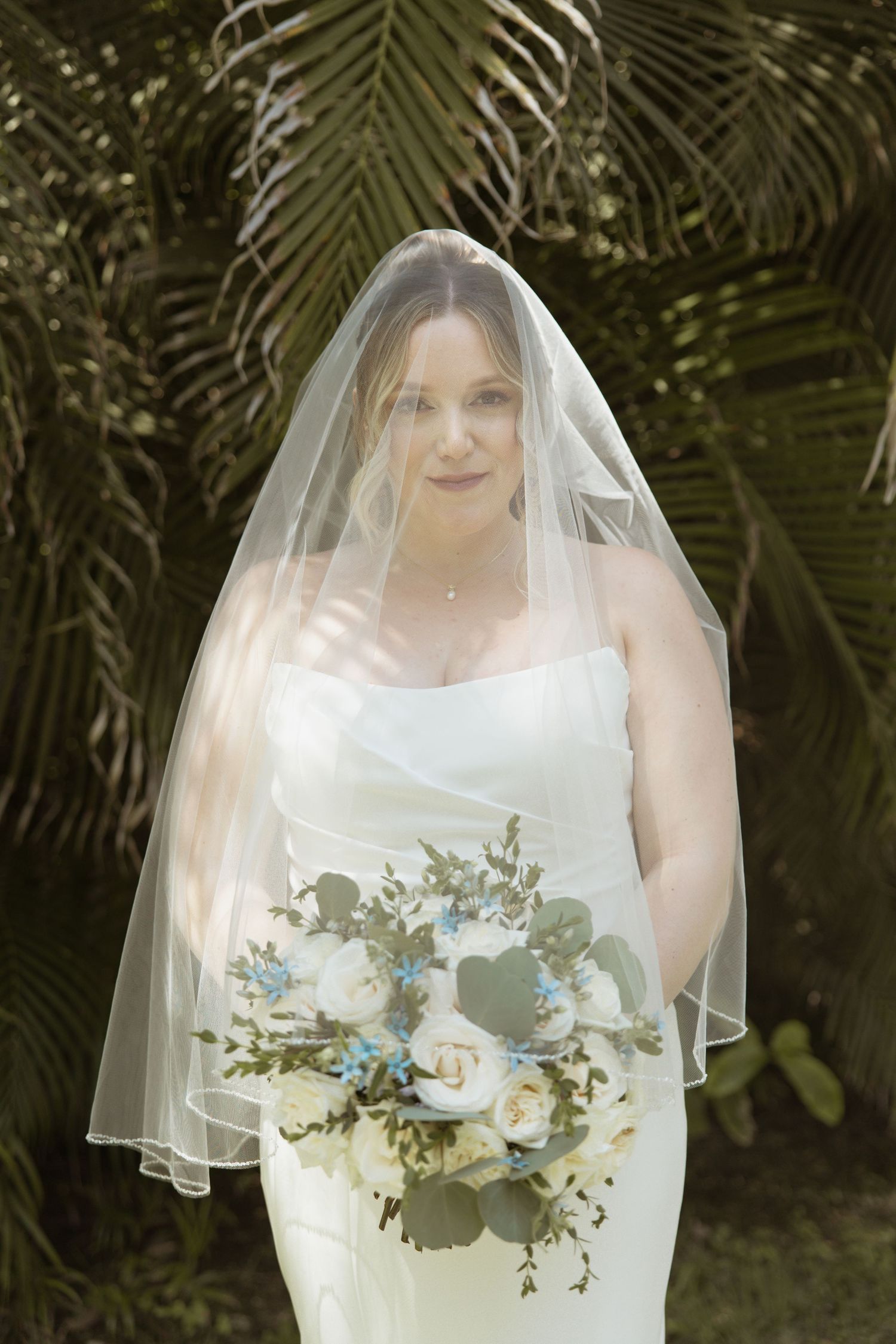 Bride in white strapless gown and veil holds bouquet of white and blue flowers with greenery outdoors.
