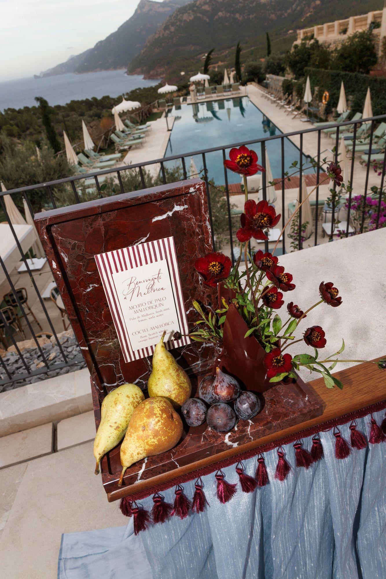 Luxury poolside setting with red flowers, pears and grapes on a wooden table overlooking a coastal mountain view. Mallorca Son Bunyola