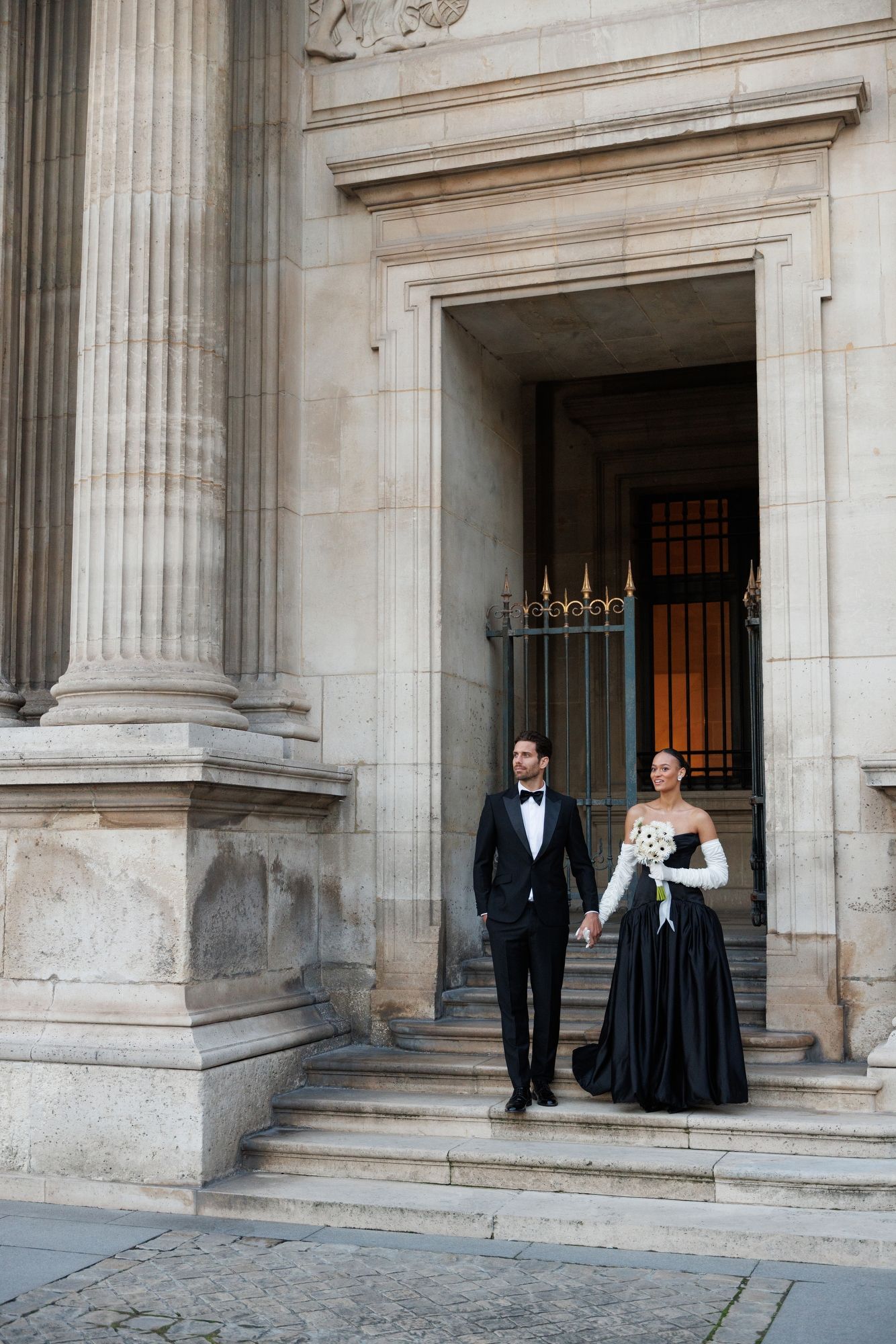Couple in formal black attire poses at grand classical building entrance with stone columns. Paris Louvre