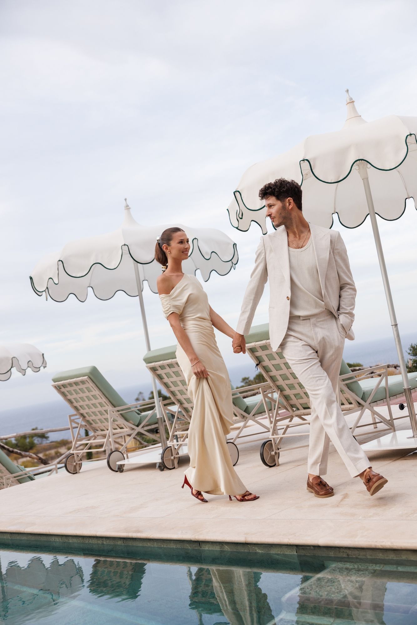 Couple in cream-colored formal wear walks playfully by poolside under white umbrellas. Mallorca Son Bunyola