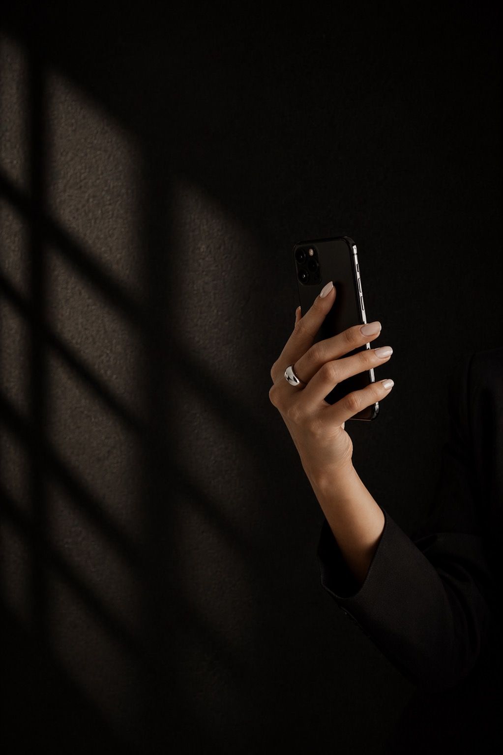 A hand wearing a silver ring holds a black smartphone against a dark wall with dramatic window light shadows.