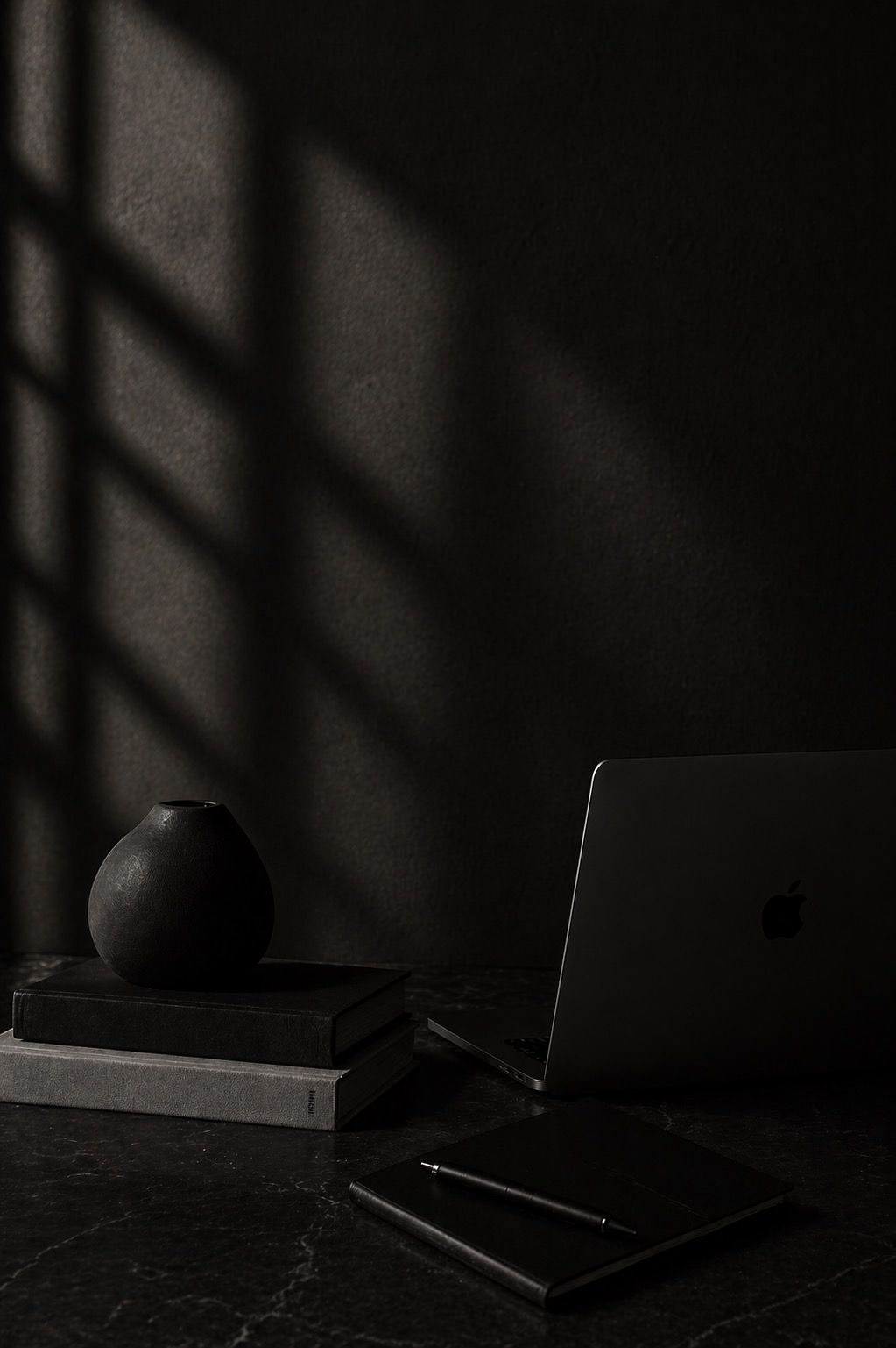 Dark minimalist workspace with MacBook, black vase on books, and dramatic window shadows on dark wall.