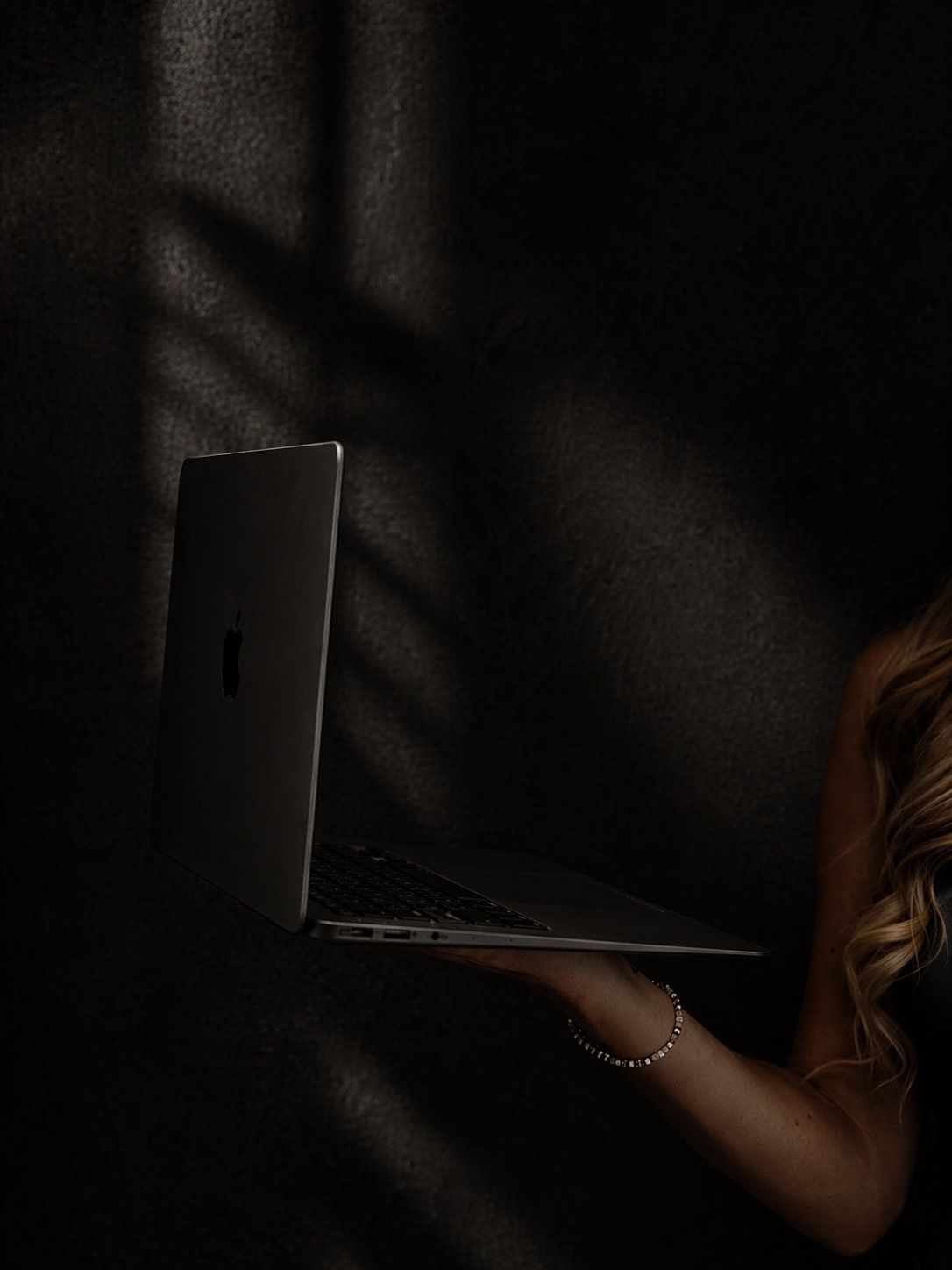 A woman with blonde hair holds an Apple MacBook laptop in a dramatic dark shadow-lit room.